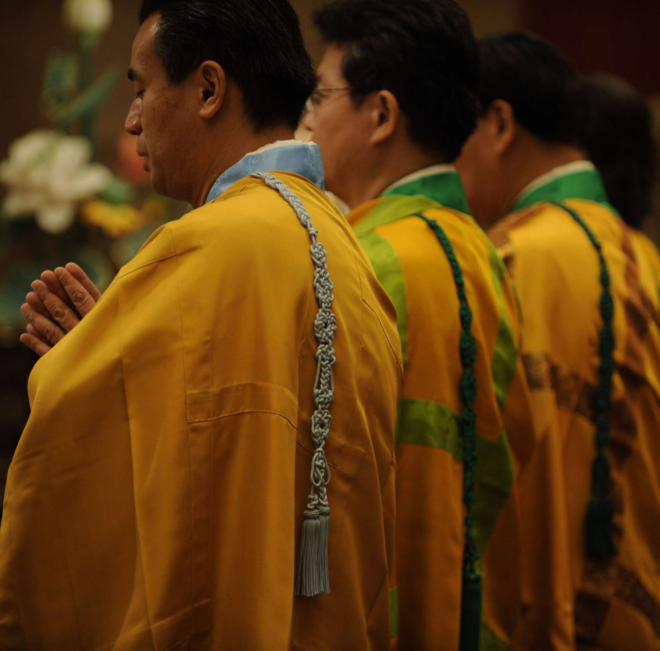 Three Buddhist priests in yellow robes, wearing green and blue kesa, and with knotted cords over their left shoulders, stand with hands folded in prayer facing left.