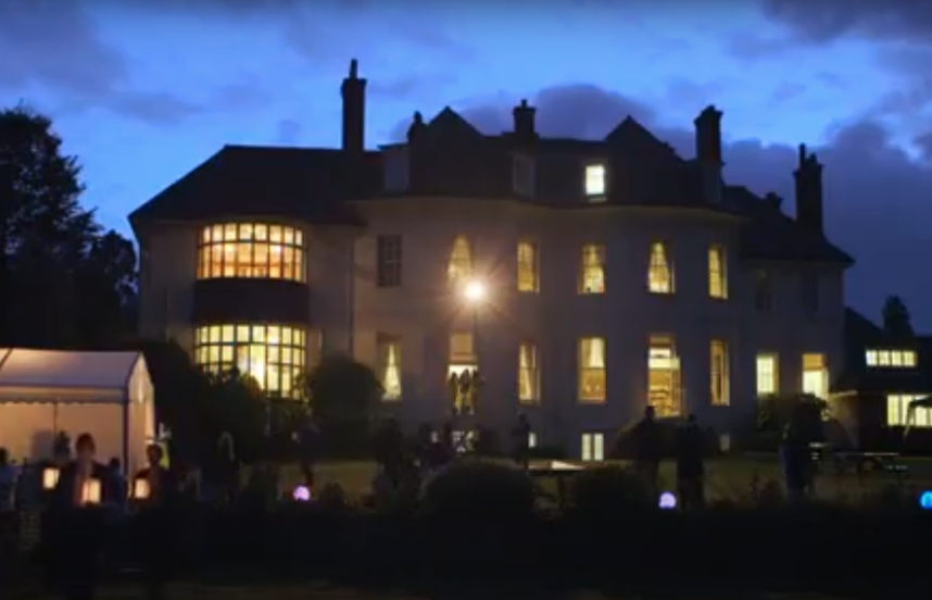 A large manor-style house stands against an evening sky with windows illuminated from within; silhouettes of people holding glowing lanterns can be seen.