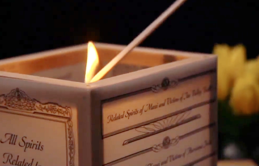 A long, lit matchstick reaches into the interior of a paper lantern to light the candle inside.