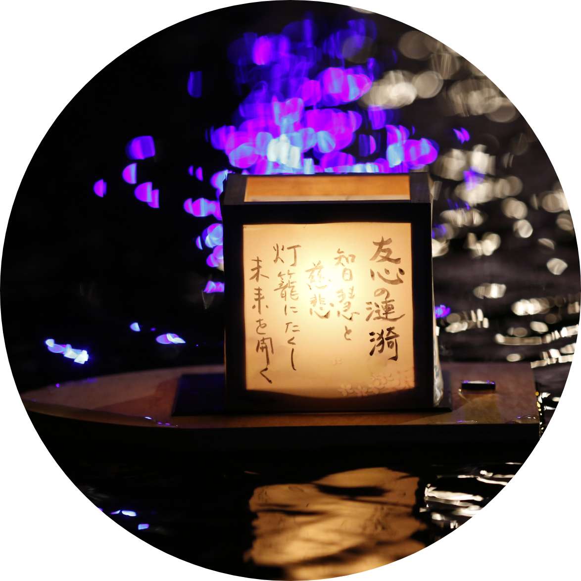 A glowing paper lantern decorated with messages in Japanese floats on water at night.