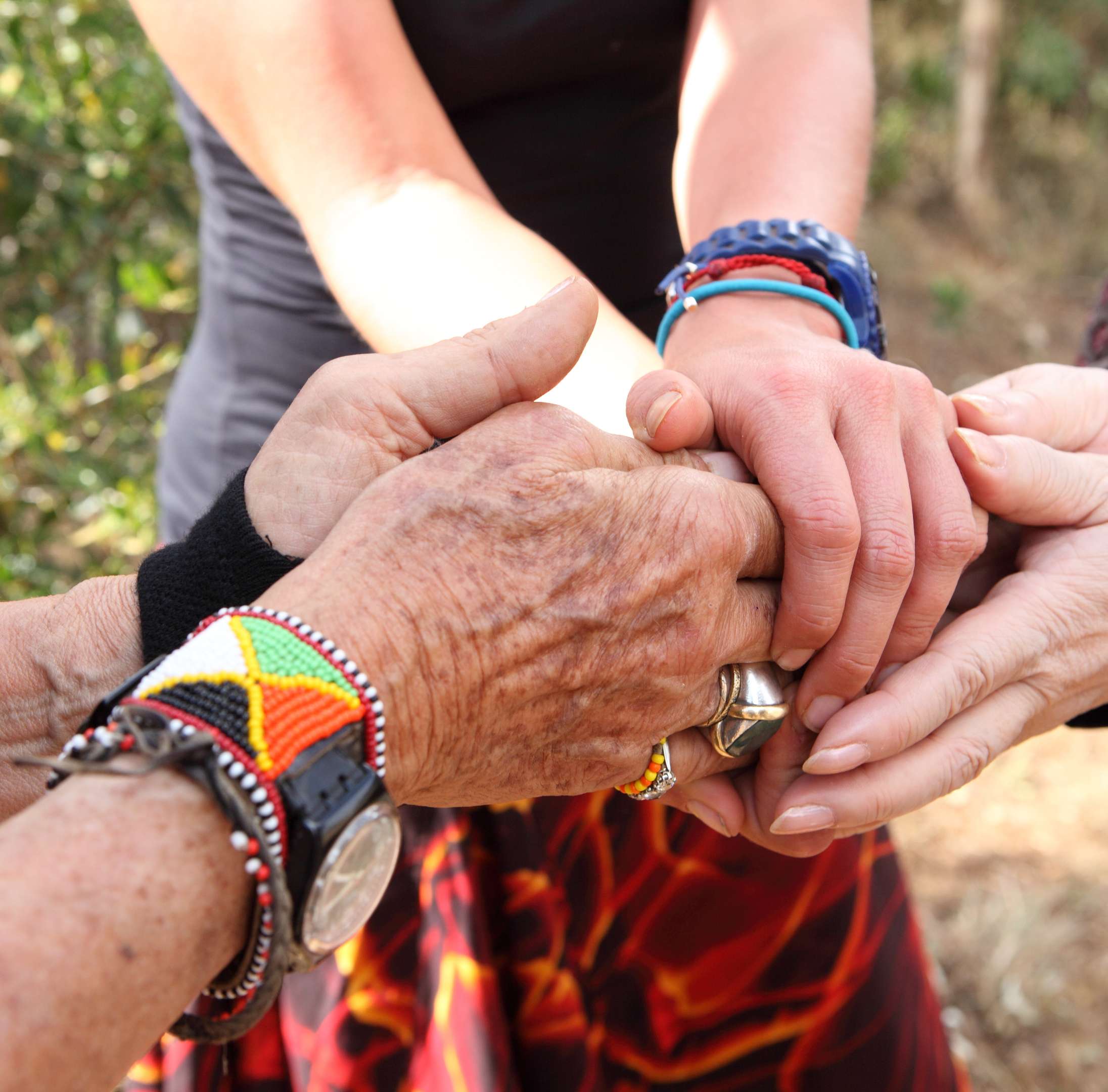 Three pairs of hands of varying ages and skin tones, two wearing colorful bracelets, reach out and embrace each other.