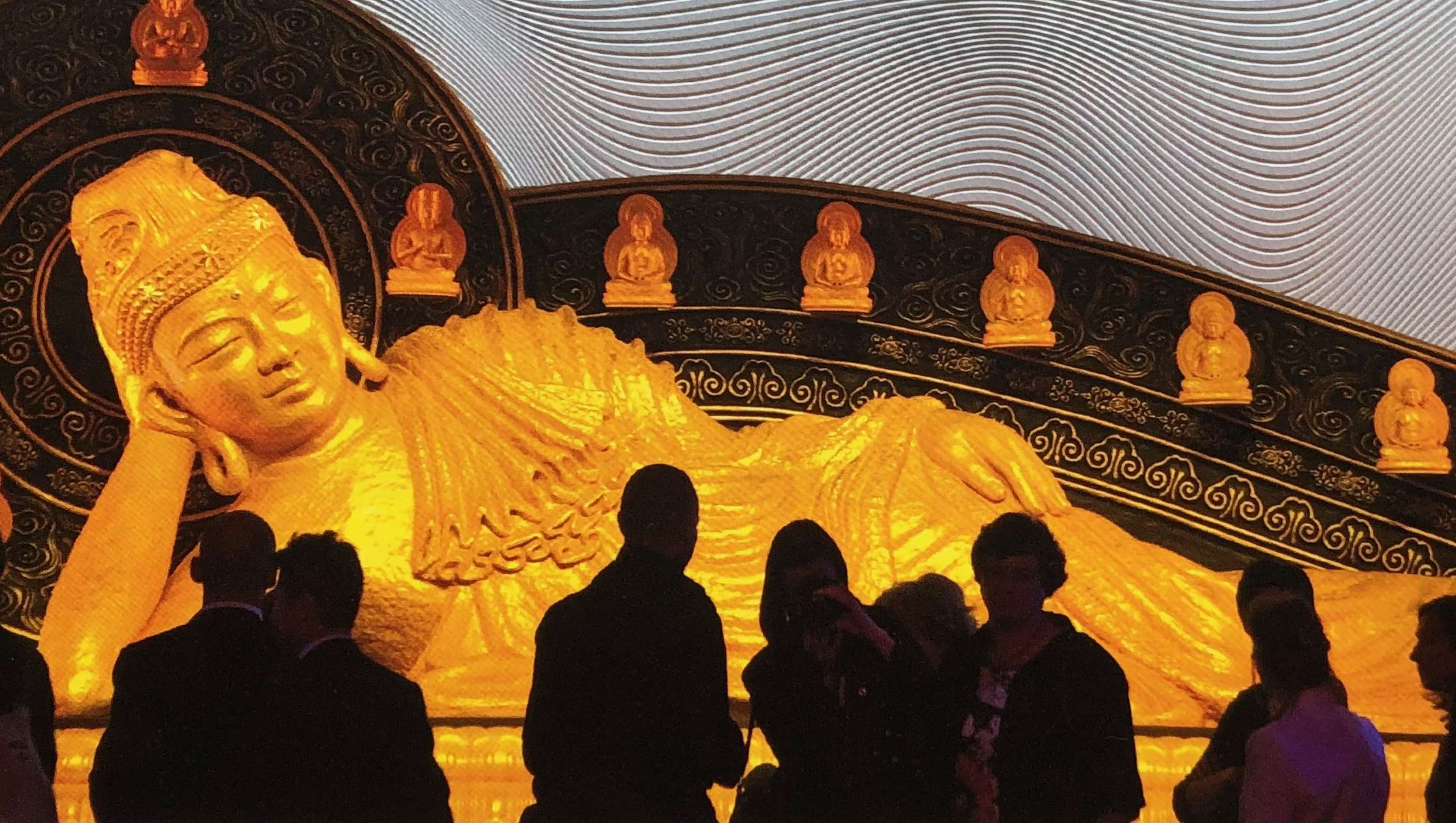A photograph of viewers silhouetted against a very large Nirvana Buddha image, in which the Buddha, reclining on his right side, resting his head on his right hand, appears to be looking at them.
