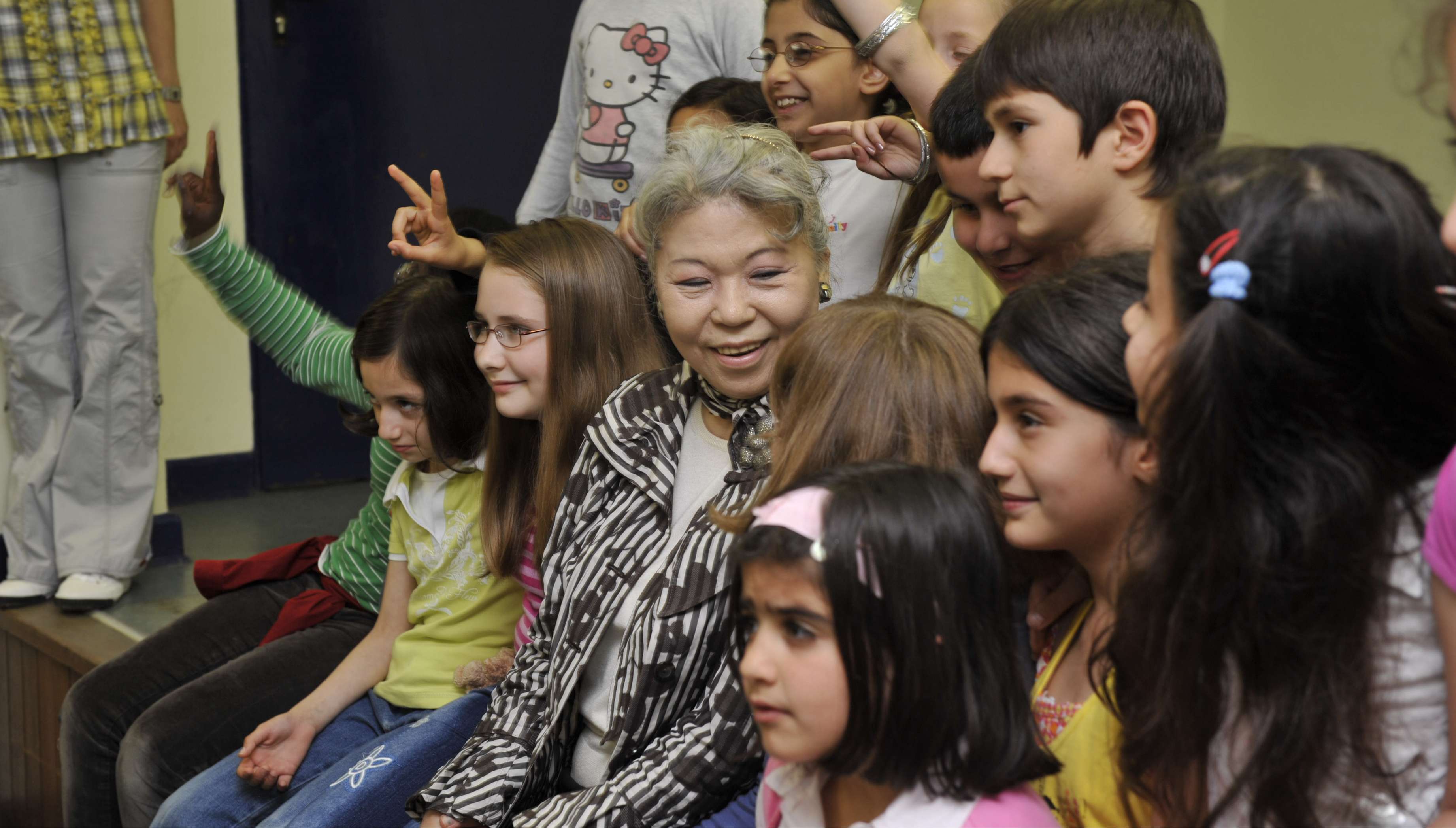 Her Holiness sits, smiling, with a diverse group of children as they participate in an activity.