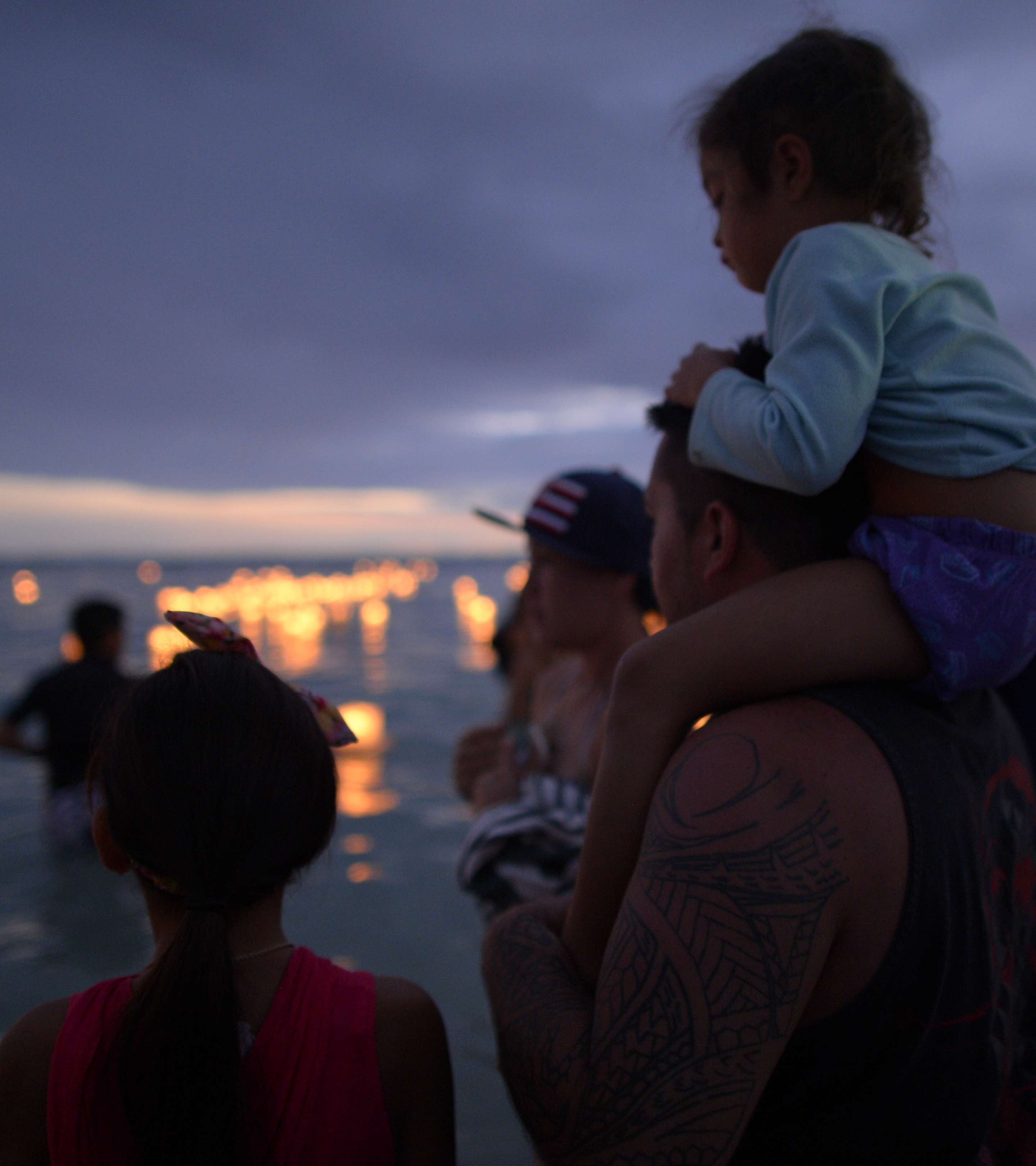 At dusk a father wading in the sea, holds a child on his shoulders as people look on at glowing lanterns floating in the distance.