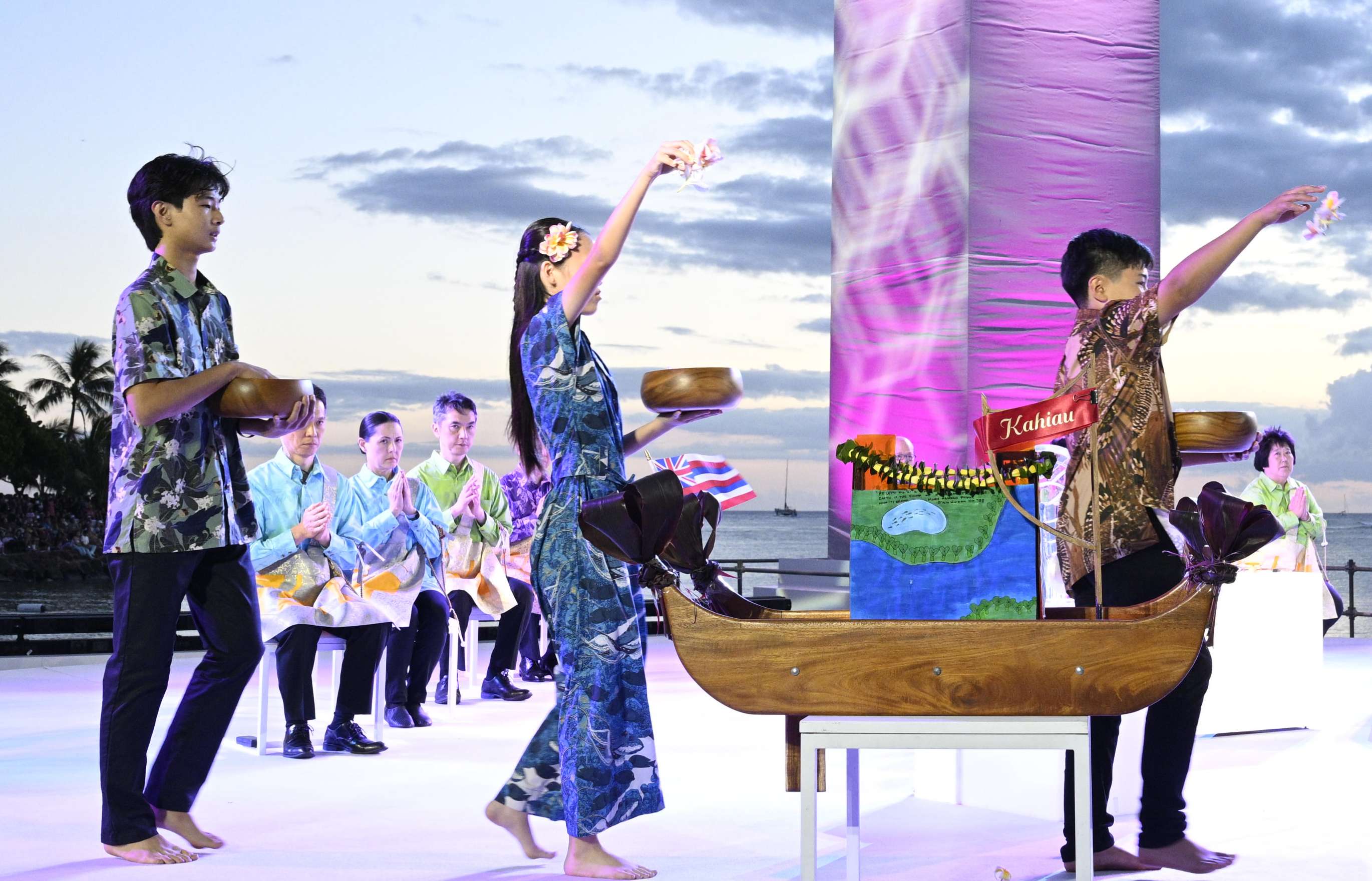 Four men and women wearing floral print clothing walk across an outdoor stage under a dusk sky, holding bowls filled with flower petals in their left hands, and strewing them from their upraised right hands; two boat-like wooden and paper lanterns sit atop plinths in the foreground; clergy members wearing blue robes, sitting with hands folded can be seen toward the back of the stage.