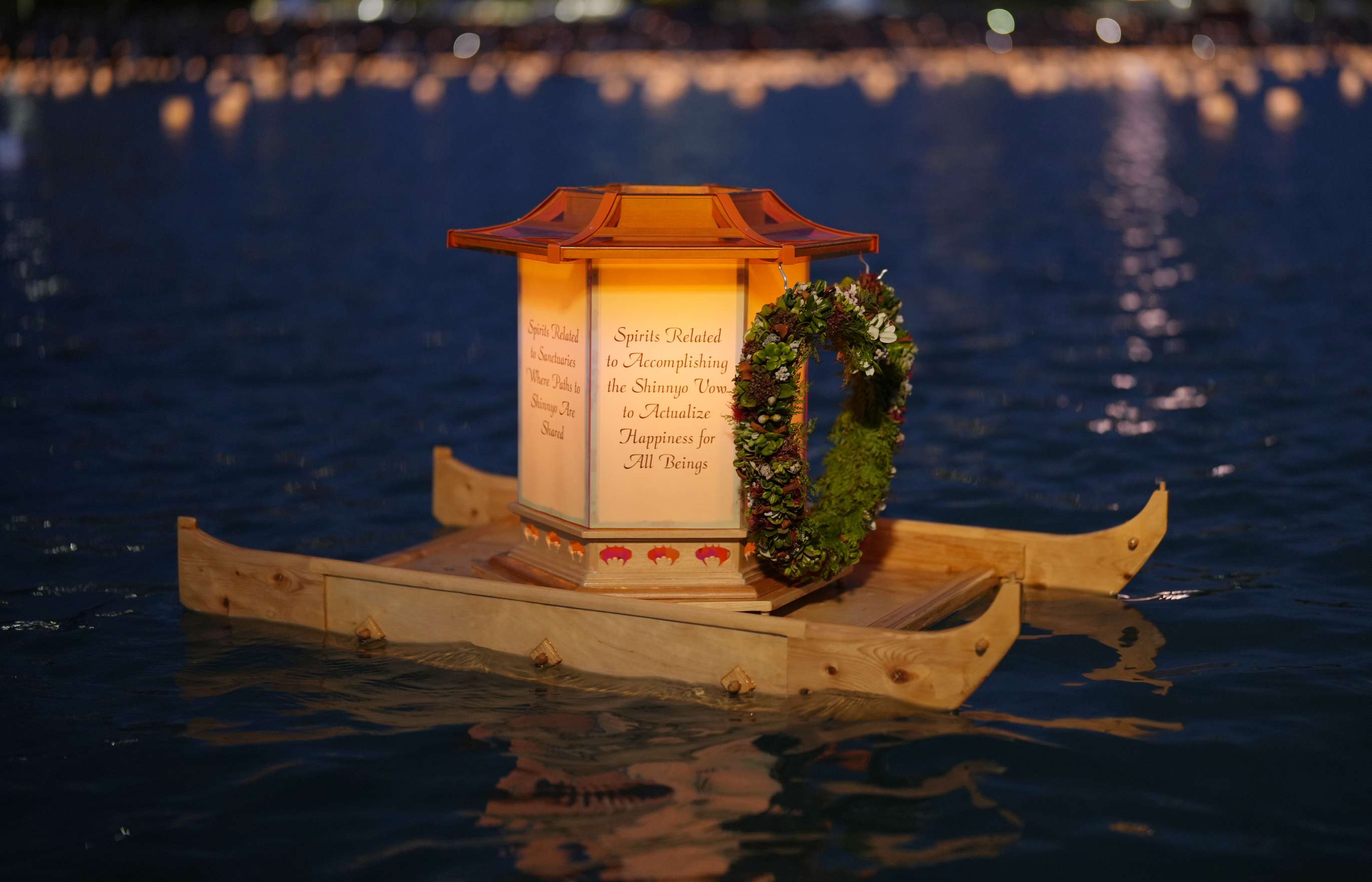 With a soft orange glow, a hexagonal paper lantern bearing prayers for hope and healing for all beings, adorned with a wreath-like lei, and fixed atop a wooden boat-like base, floats in the night time waters of a bay with a view of other floating lanterns and city lights and buildings in the deep background.