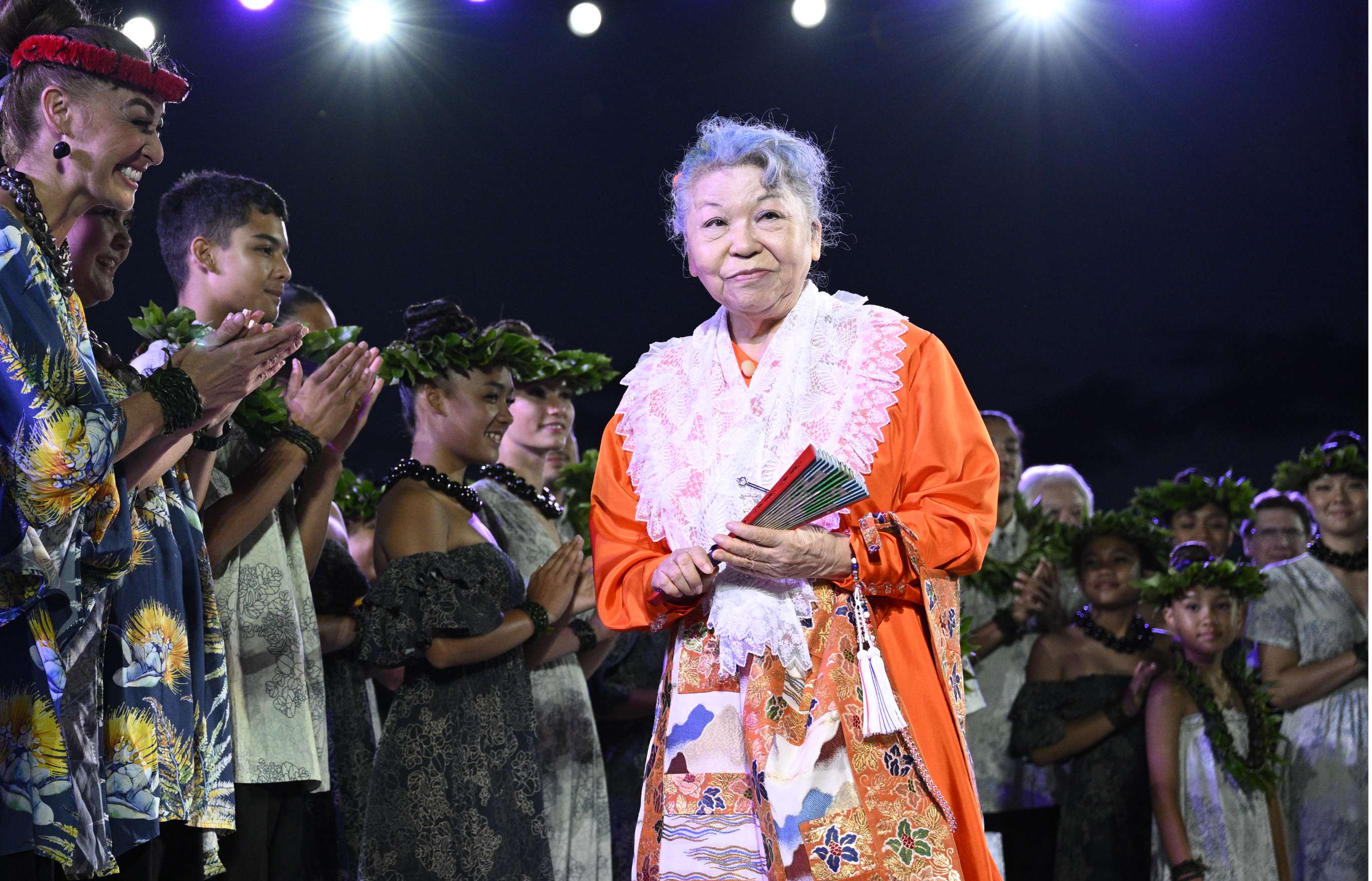 Beneath stage lights and a night sky, a grey-haired woman in bright orange robes with a lace shawl and brocade apron, holds a folding fan in her hands and smiles warmly as a large group of singers dressed in floral print clothing cheerfully smile at her.