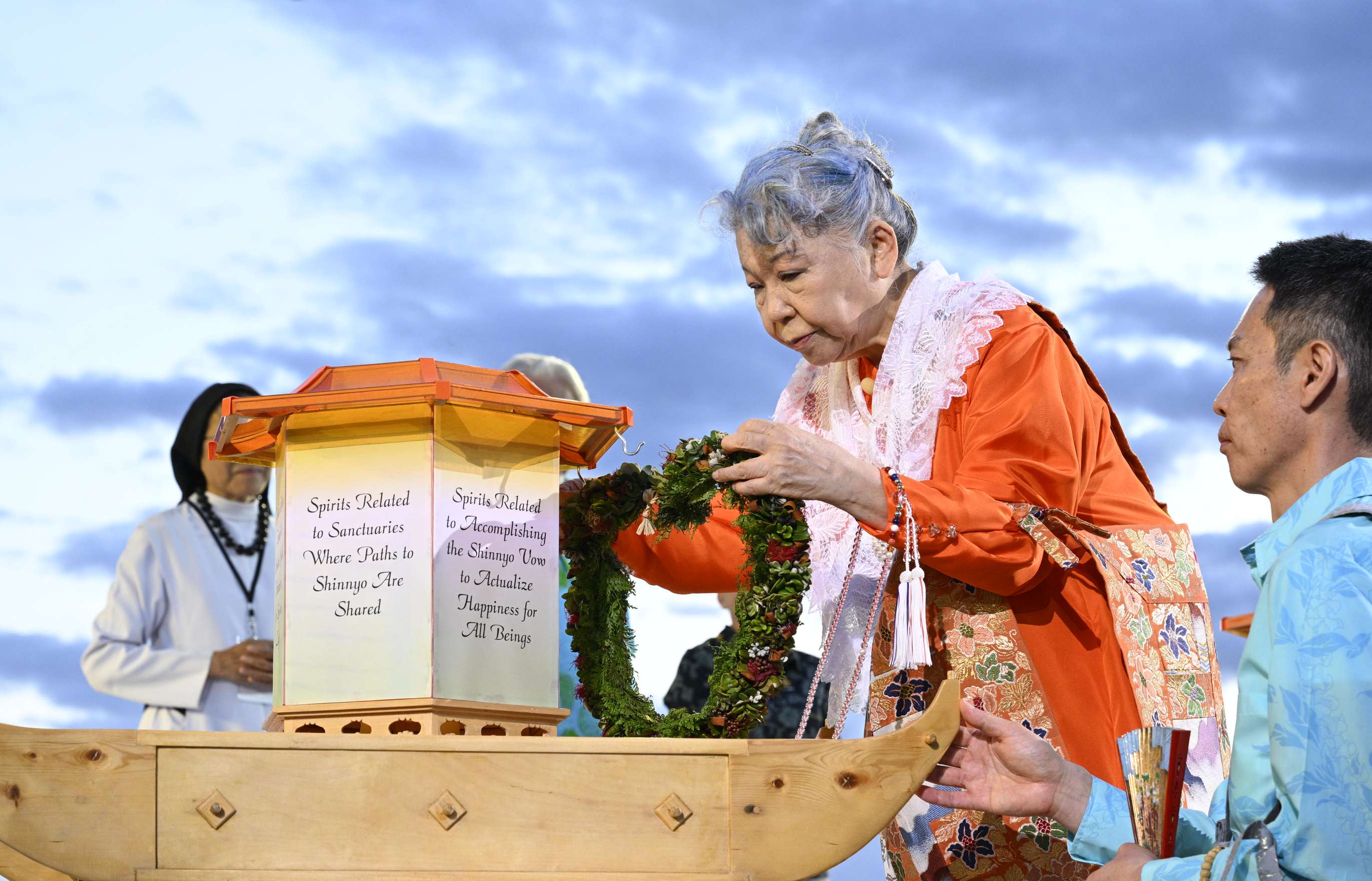 Against the backdrop of a dusk sky, an elder, grey-haired woman wearing bright orange robes and a white lace shawl bows slightly as she places a garlanded wreath onto a boat-like wooden and paper lantern; an attendant wearing a blue shirt, holding a folding fan, and prayer beads kneels to her left, steadying the lantern.