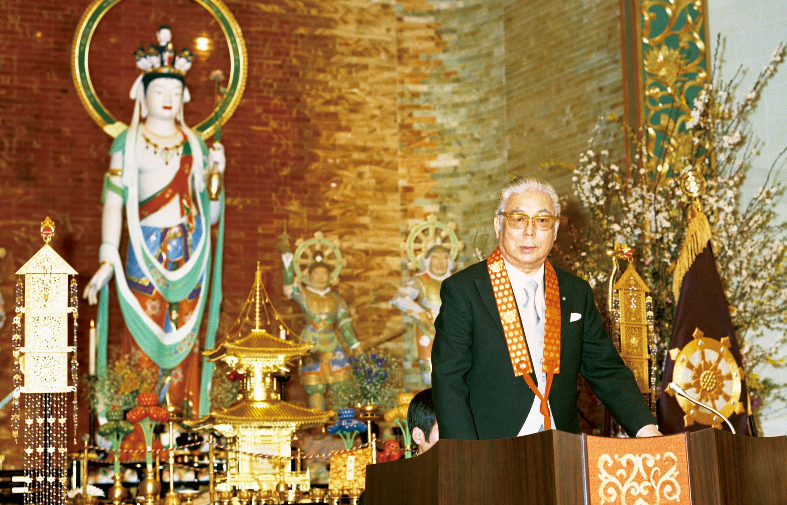 A white haired Japanese man, dressed in a black suit jacket and tie with a vermillion surplice around his neck stands, speaking at a podium decorated with ornate filigree in front of a gold-tiled knave housing a large statue of a standing bodhisattva dressed in flowing scarves and silken garments; a finely crafted altar with a golden pagoda is visible in front of the statue.