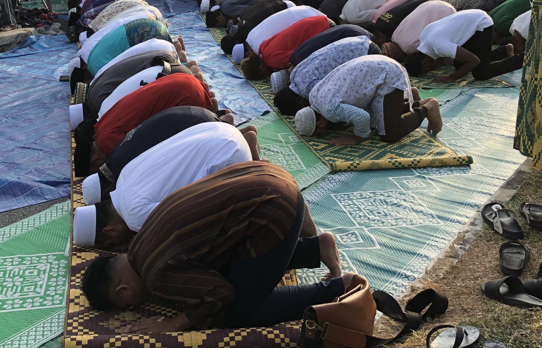 Rows of men wearing long shirts and skull caps bowing on prayer carpets with their heads, hands, and feet touching the ground; discarded sandals can be seen nearby.