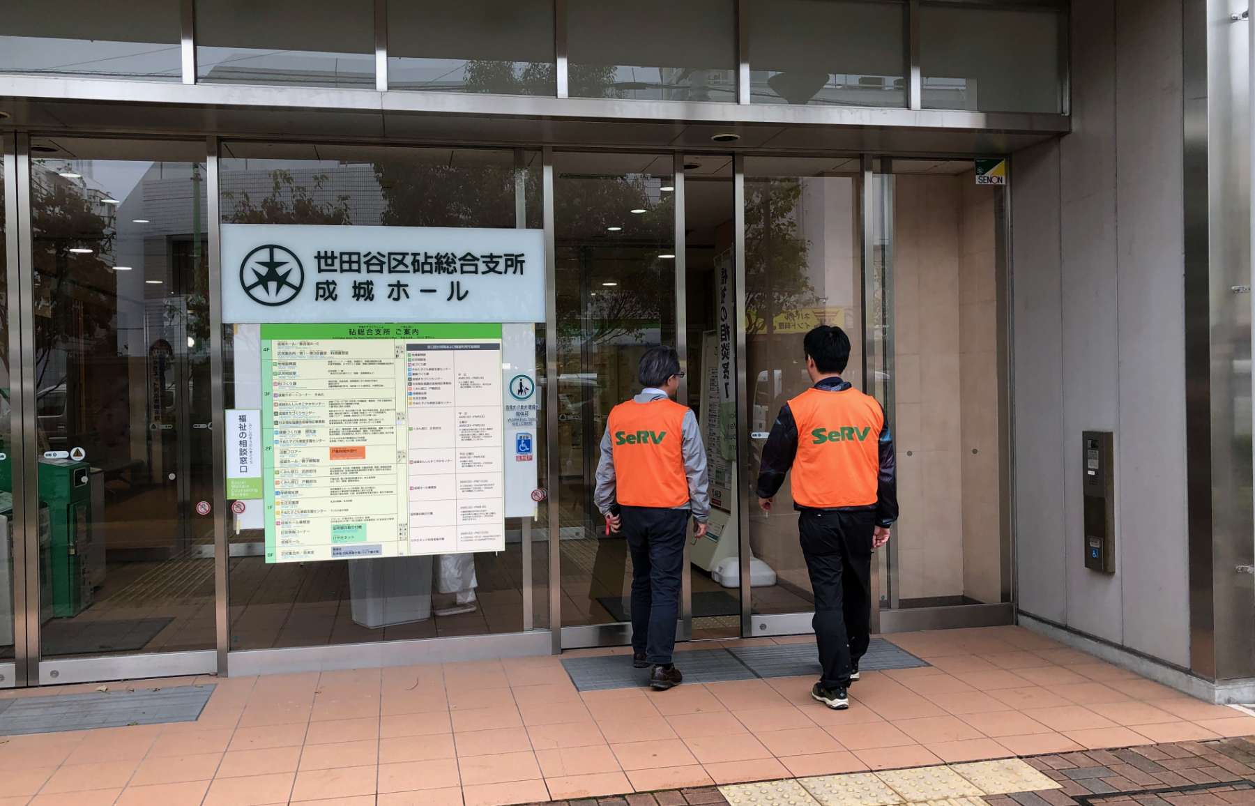 Two men wearing orange SeRV volunteer vests over suits approach the glass doors at the entry to a building with a large sign in Japanese next to the door.