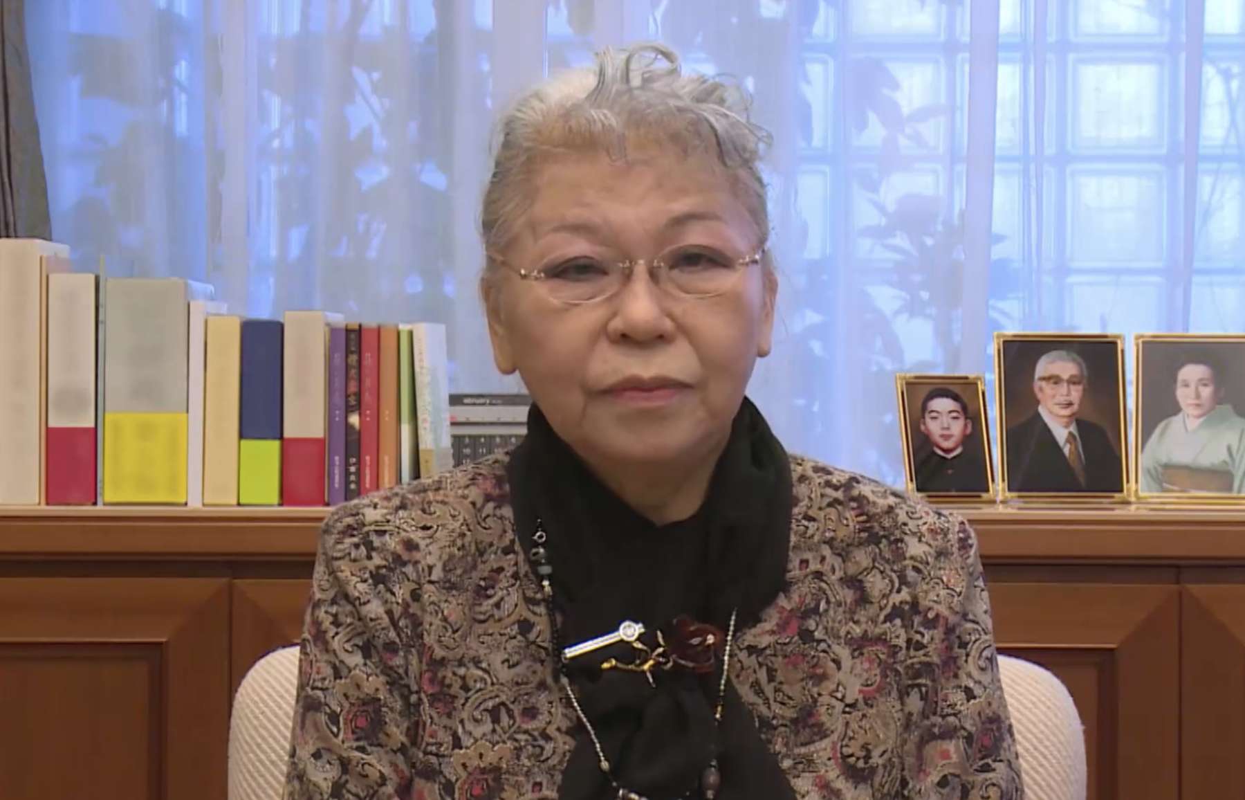 Her Holiness Shinso Ito, wearing spectacles, a patterned blouse with a black scarf, sitting in front of a window with a shelf displaying photos, looks directly into the camera.