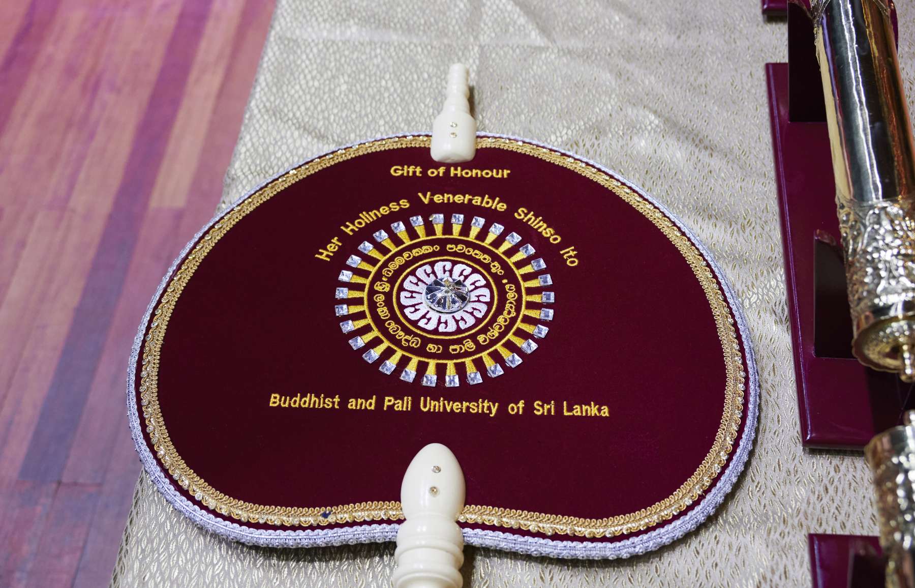 A maroon ceremonial hand-held fan decorated with a circular pattern, Singala letters, and the words, “Gift of Honour, Her Holiness Shinso Ito, Buddhist and Pali University of Sri Lanka.”