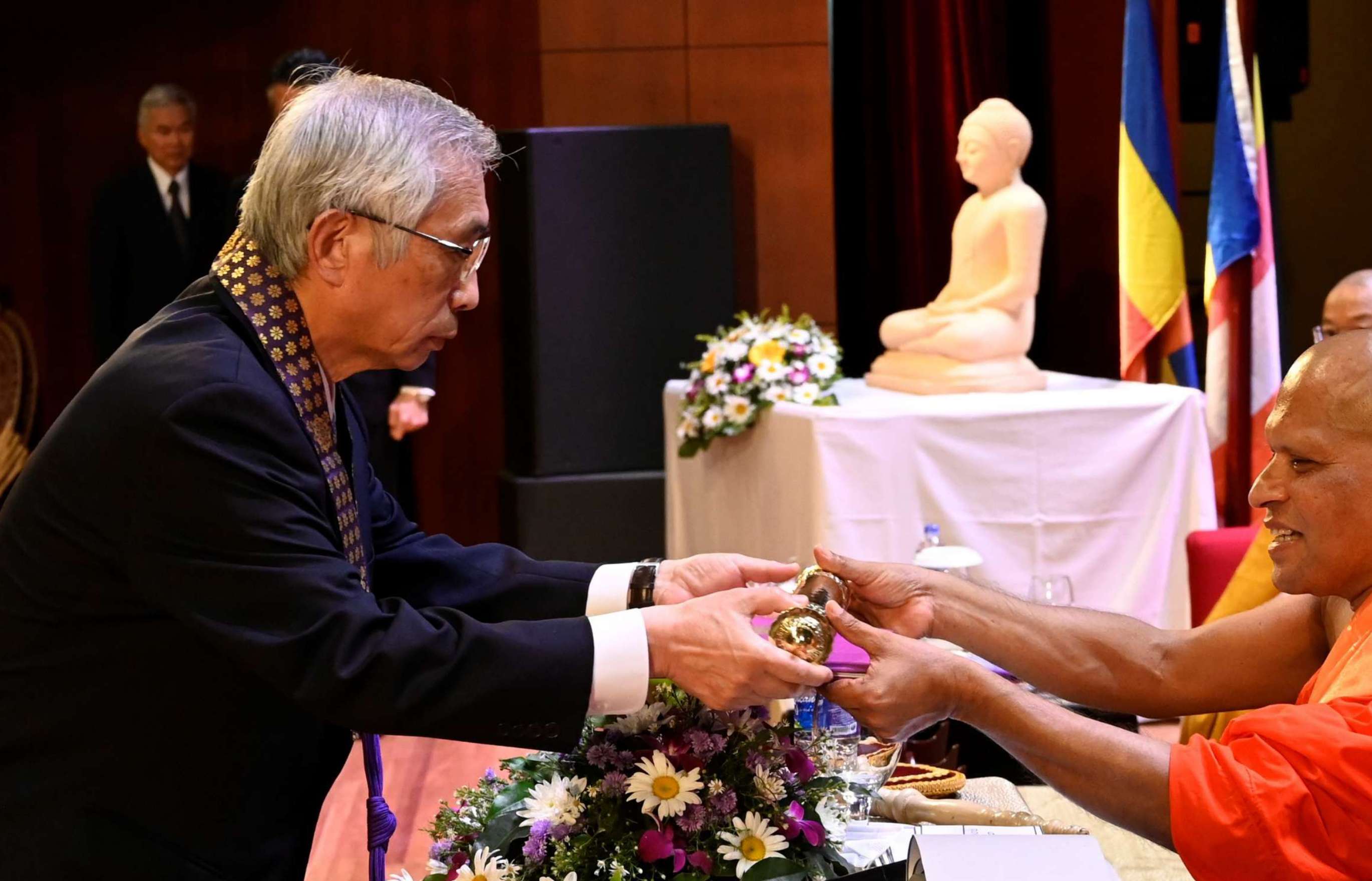 A standing gray-haired man in a suit, wearing a kesa around his neck, accepts a golden cylinder from a seated shaven-headed monk in orange robes; a statue of Buddha can be seen in the background.