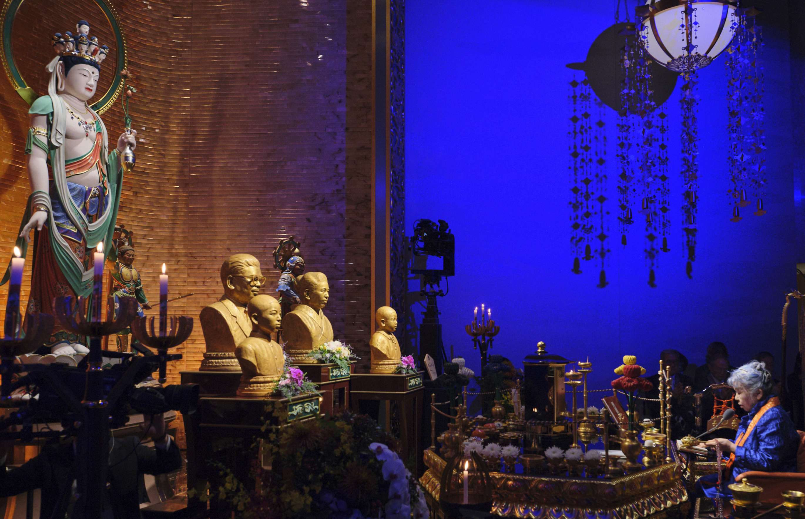 A gray-haired Japanese woman in blue dress with an orange surplice around her neck, seated before an ornate altar on which are arranged busts and a large standing statue of a bodhisattva, holds a string of prayer beads and a censer as she seems intently focused on conducting a rite; the altar backdrop made of gold-colored bricks glows warmly in the space which is otherwise bathed in deep blue light.