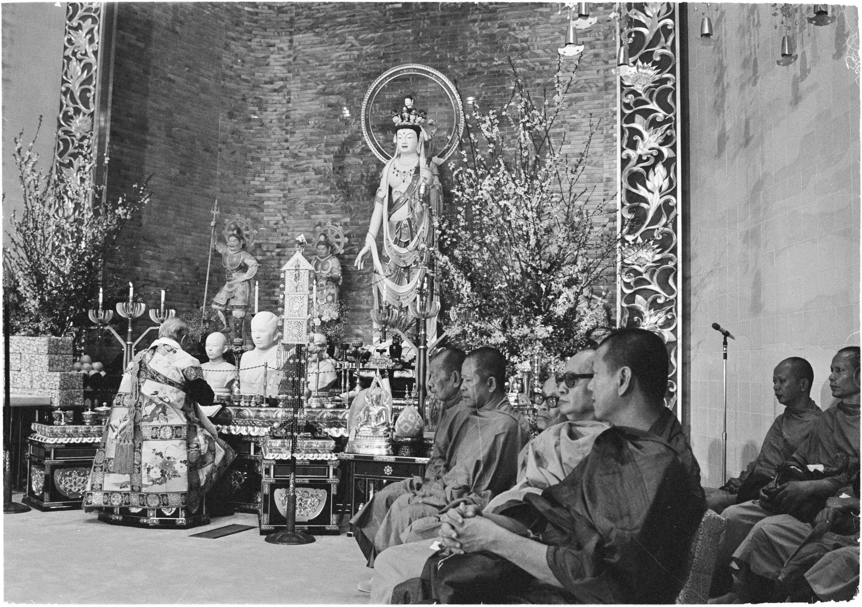 A gray-haired man dressed in brocade priestly robes sits with his back to the camera, hands folded in prayer before an ornate altar decorated with numerous ritual items. He faces a large statue of a standing bodhisattva in a tall temple nave; a row of monks in robes seated in chairs, watching the proceedings, can be seen in the right-hand corner of the image.