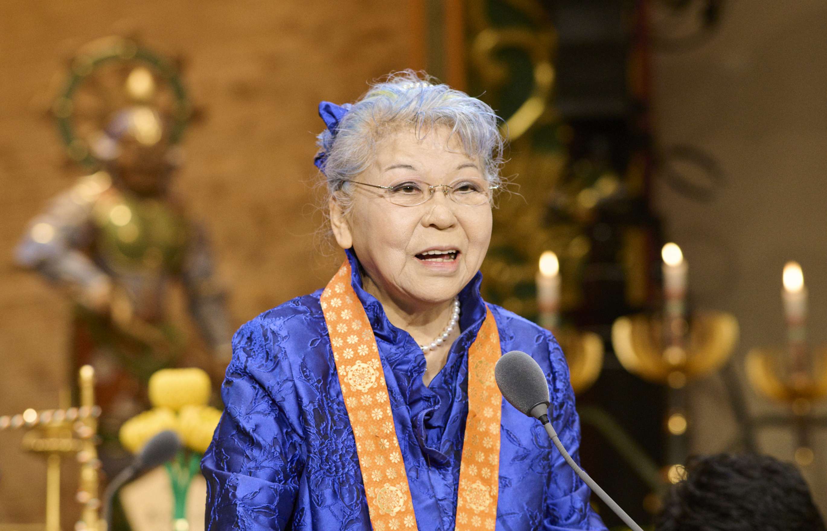 A gray-haired Japanese woman wearing a blue dress with a vermillion surplice around her neck stands at a podium, smiling, delivering an address; a golden hued altar backdrop and standing statue can be seen in the background.
