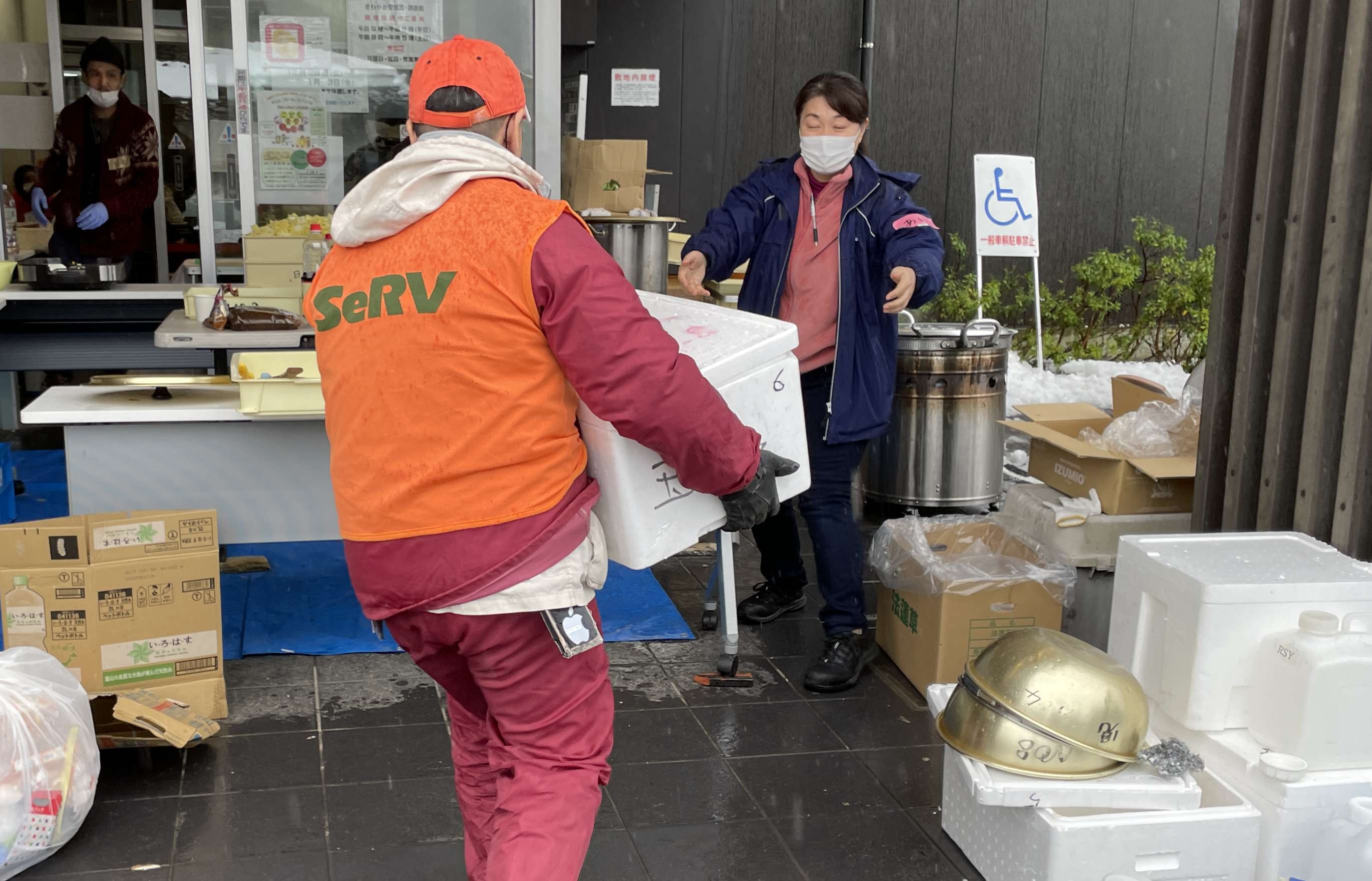 Two men wearing medical masks and wearing orange hi-vis vests with the SeRV logo on them deliver boxes of supplies to a makeshift station for distribution of relief supplies set up atop blue tarps in the entrance to a building; tiled, high peaked Japanese rooftops are visible in the background.