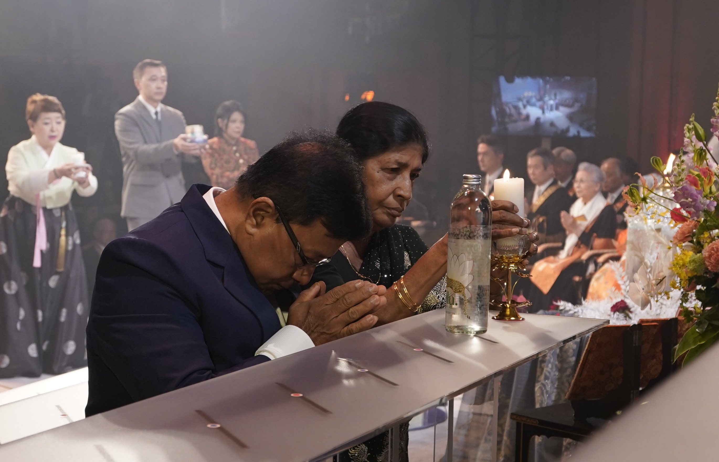 A man bows, hands folded as a woman next to him reverently place a lit candle next to a bottle of water on a shrine-like platform; in the background others can be seen carrying offerings of water and fire as they wait their turn to place them on the shrine; seated elders in ceremonial robes sit with hands folded watching the procession.