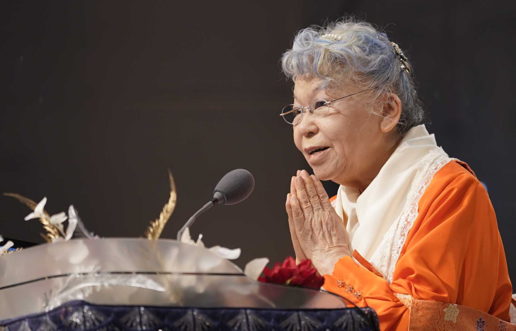 An elder Japanese woman wearing bright orange robes and a white shawl sits at a table speaking before a microphone with her hands folded in a gesture of gratitude.