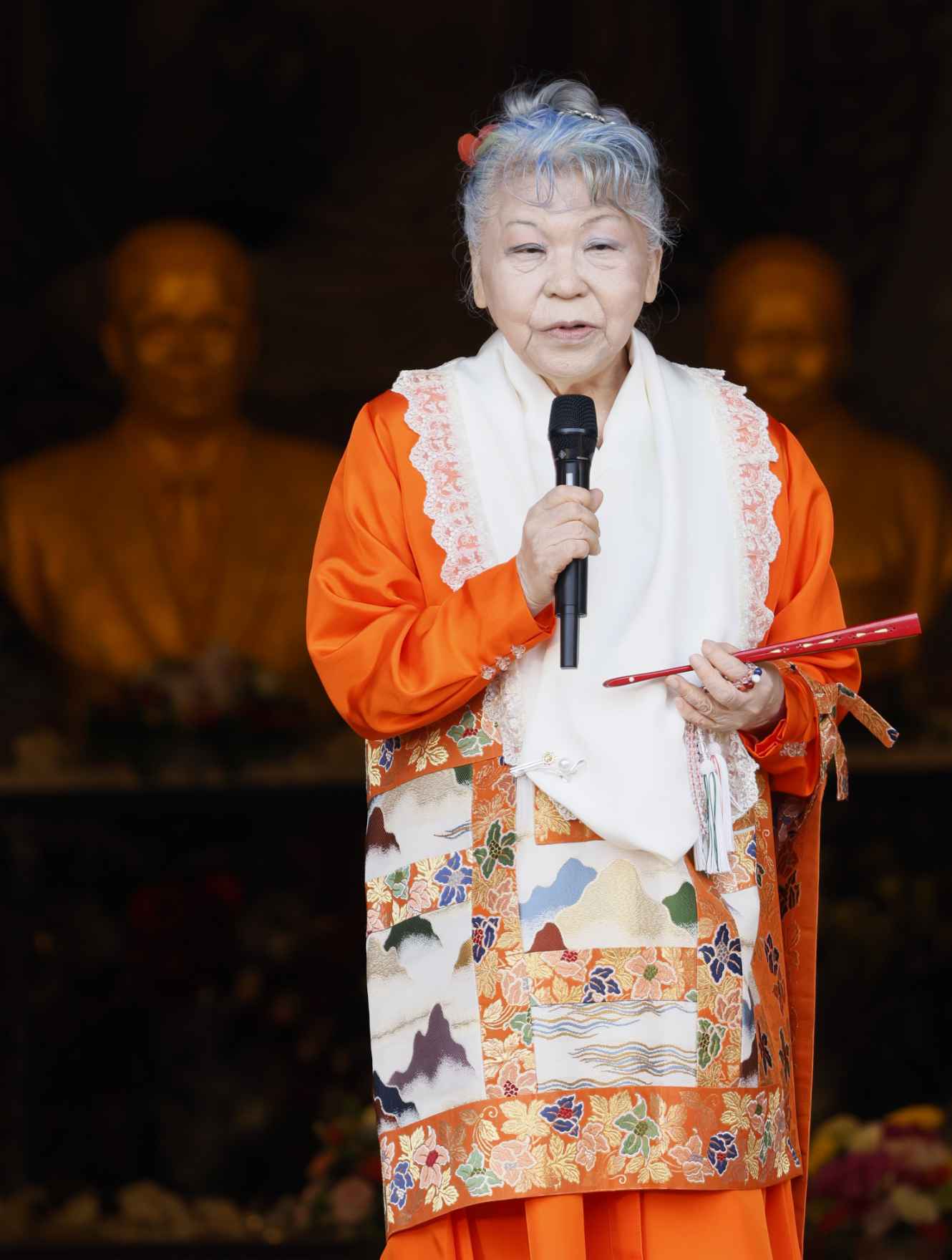 An elder Japanese woman wearing bright orange ceremonial robes with ornate brocade trim and a white lace shawl stands speaking into a microphone she holds in her right hand; she holds a folded fan in her left hand; golden hued busts can be seen in the darkened interior of the temple behind her.