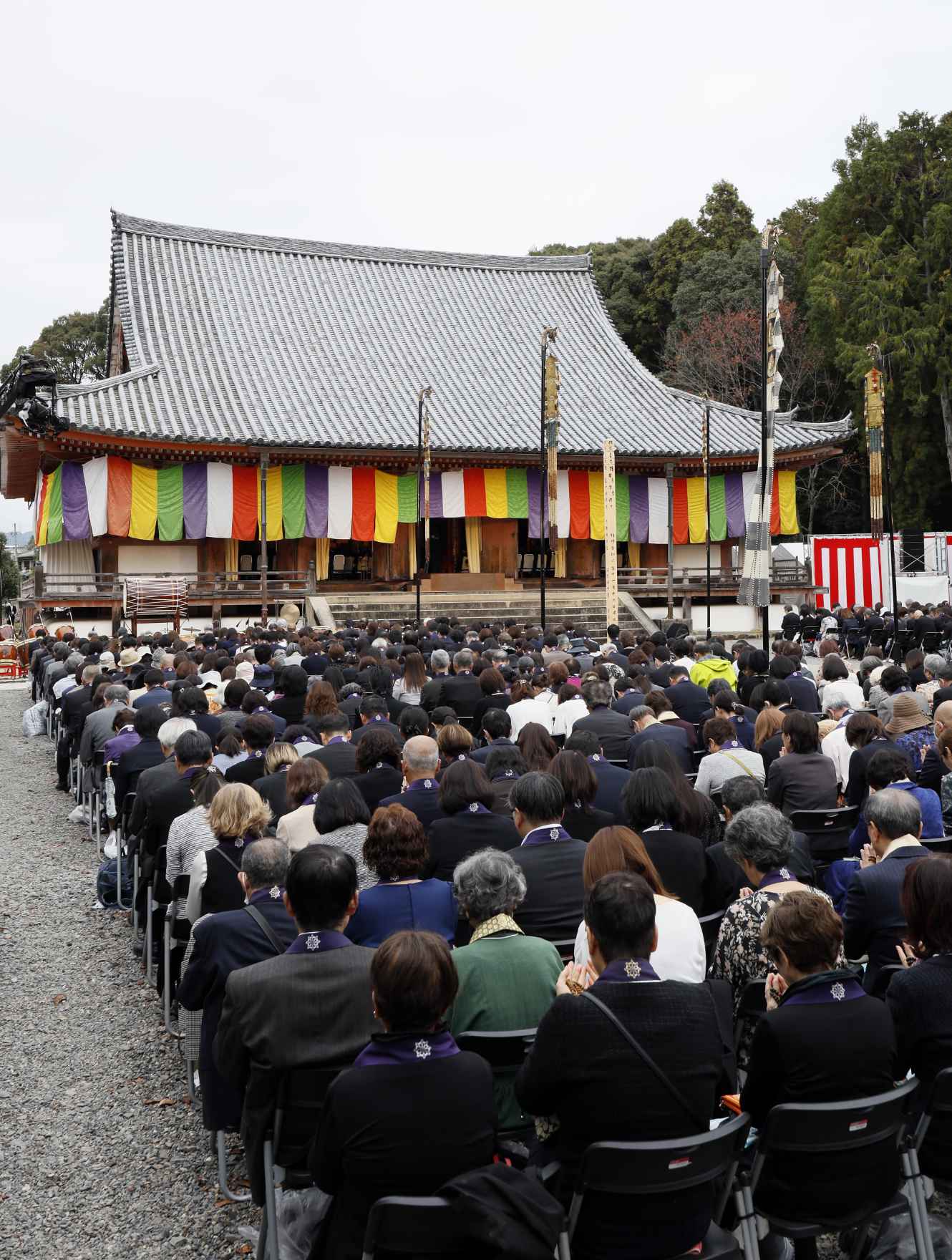 A large group of people sit in rows of chairs in the cobblestone courtyard outside of an old temple with a high-peaked roof, the eaves of which are decorated with hanging curtains, alternating white, red, yellow, green, and blue, against a backdrop of green trees and pale open sky.