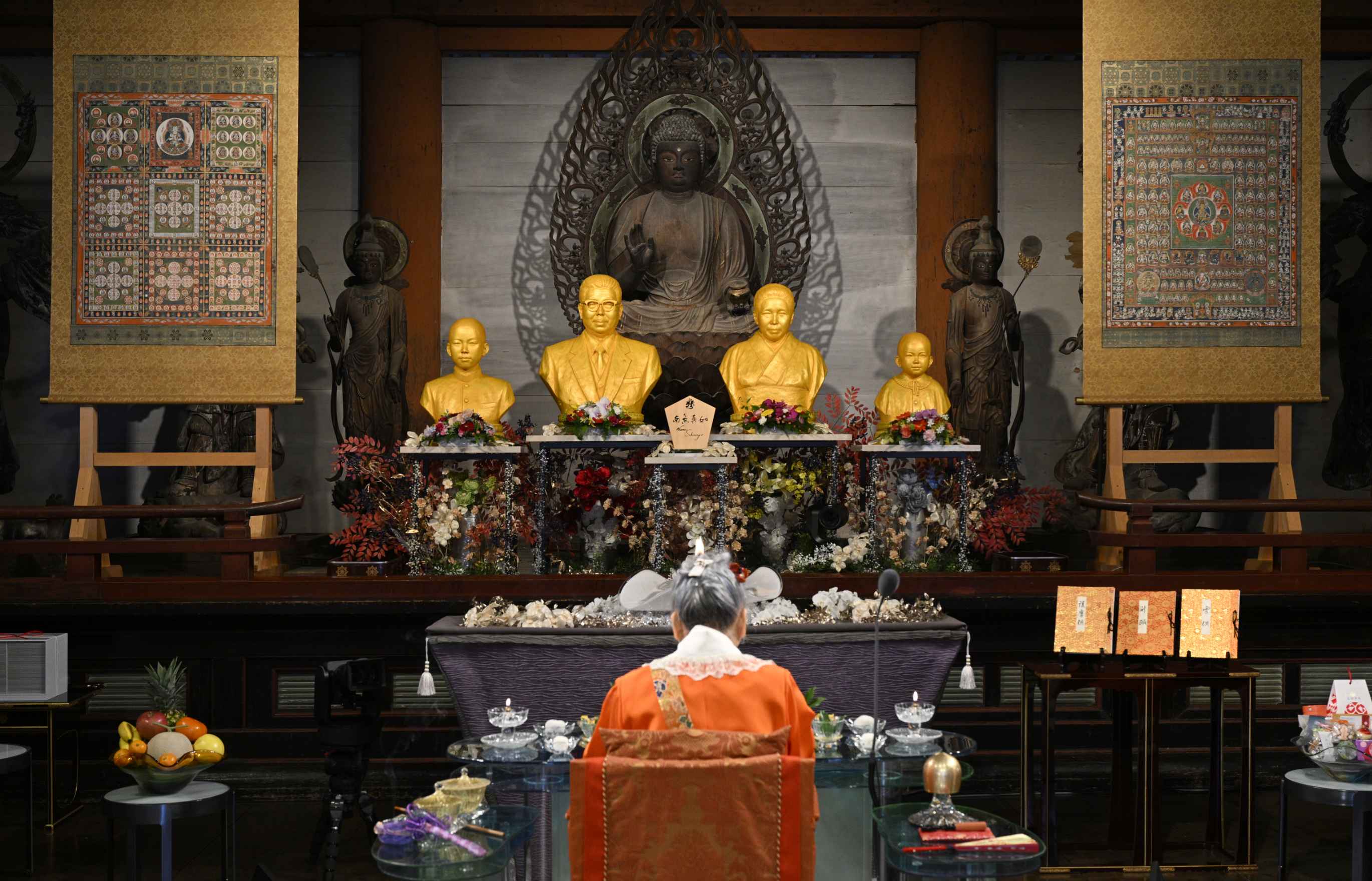  An elder Japanese woman in bright orange robes sits performing a rite with her back to the camera before a table laden with ritual offering substances, facing a large, ornately hand-carved wooden Buddha statue, in front of which four golden-hued busts representing the Shinnyo Masters and their sons have been placed; large scrolls with detailed mandalas hang to the left and right of the altar.