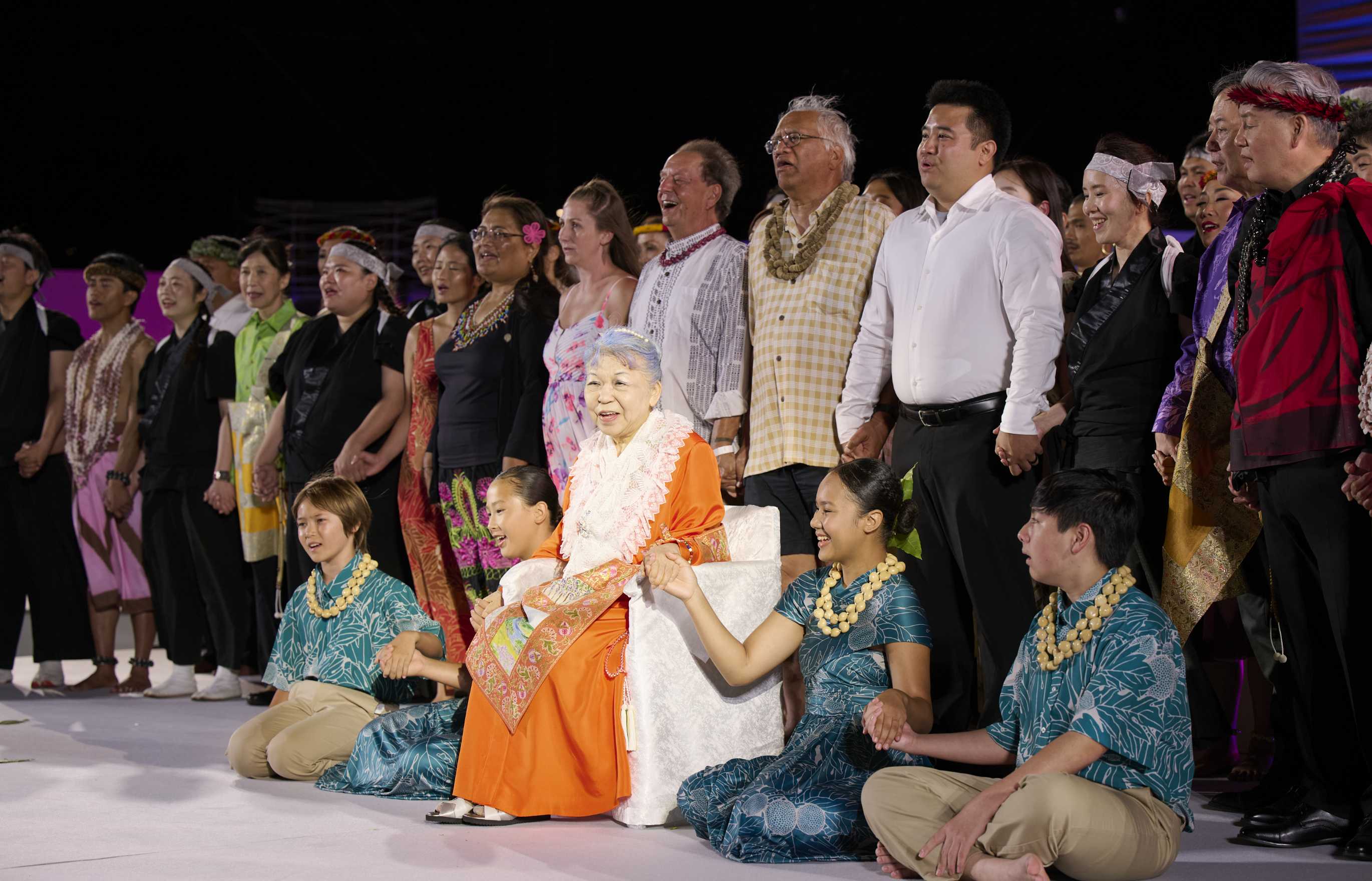 Beneath stage lights and a night sky, a grey-haired woman in bright orange robes with a lace shawl and brocade apron, sits on a chair under lights on a stage, holding hands with children seated to her left and right, smiling warmly as a large group of singers stand in a row behind her on the stage singing.