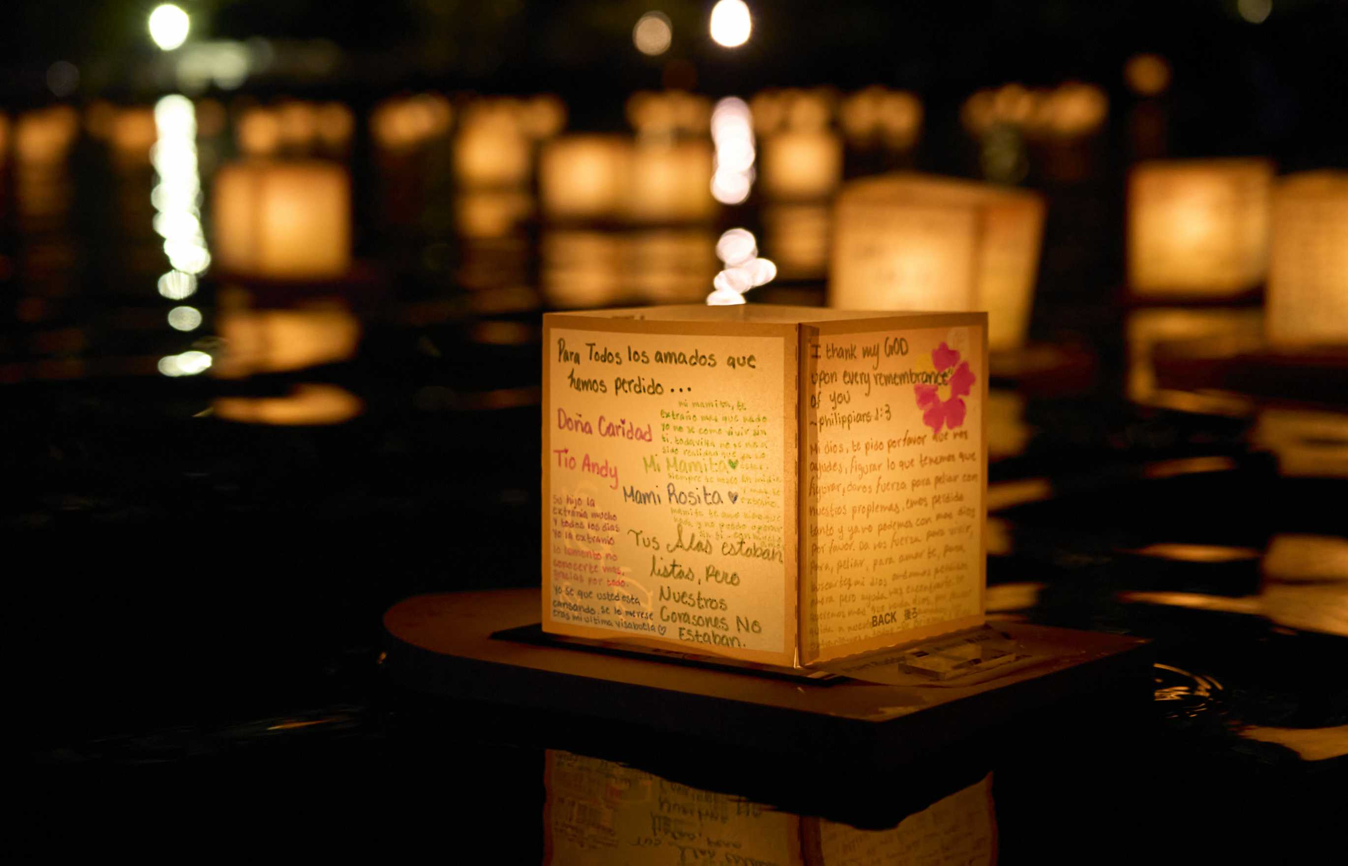 An intricately decorated square lantern, with writing in different colors, hands, and languages, emits a soft orange glow as it floats in the night time waters of a bay with a view of other floating lanterns and shorelights in the deep background.
