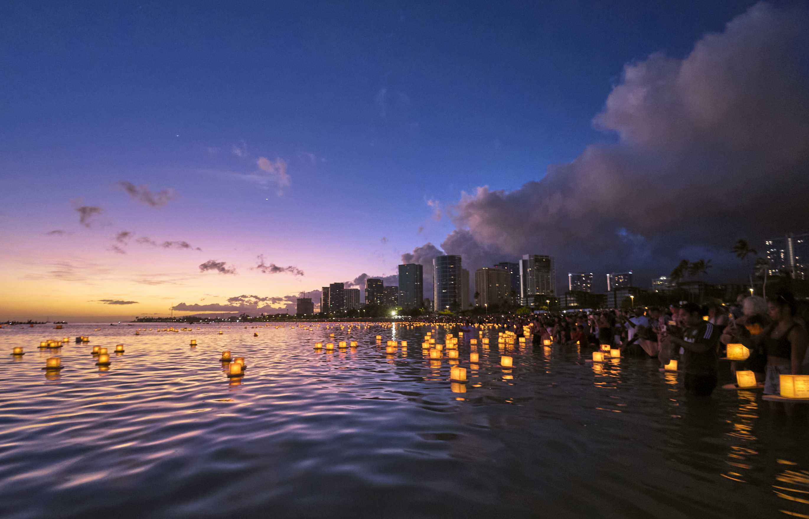 Glowing lanterns are released into the calm ocean waters of a bay at dusk; the gentle glow of a fading sunset and the Honolulu skyline are visible in the background; the orange glow of the lantern and of the glimmer of sunset are reflected in the ripples of the water.