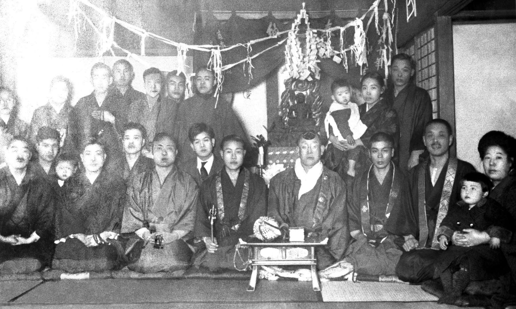 A group portrait in a temple space in which Shinjo, wearing Dharma robes, sits to the right of the officiating priest, Tomoji stands behind them holding infant Chibun.