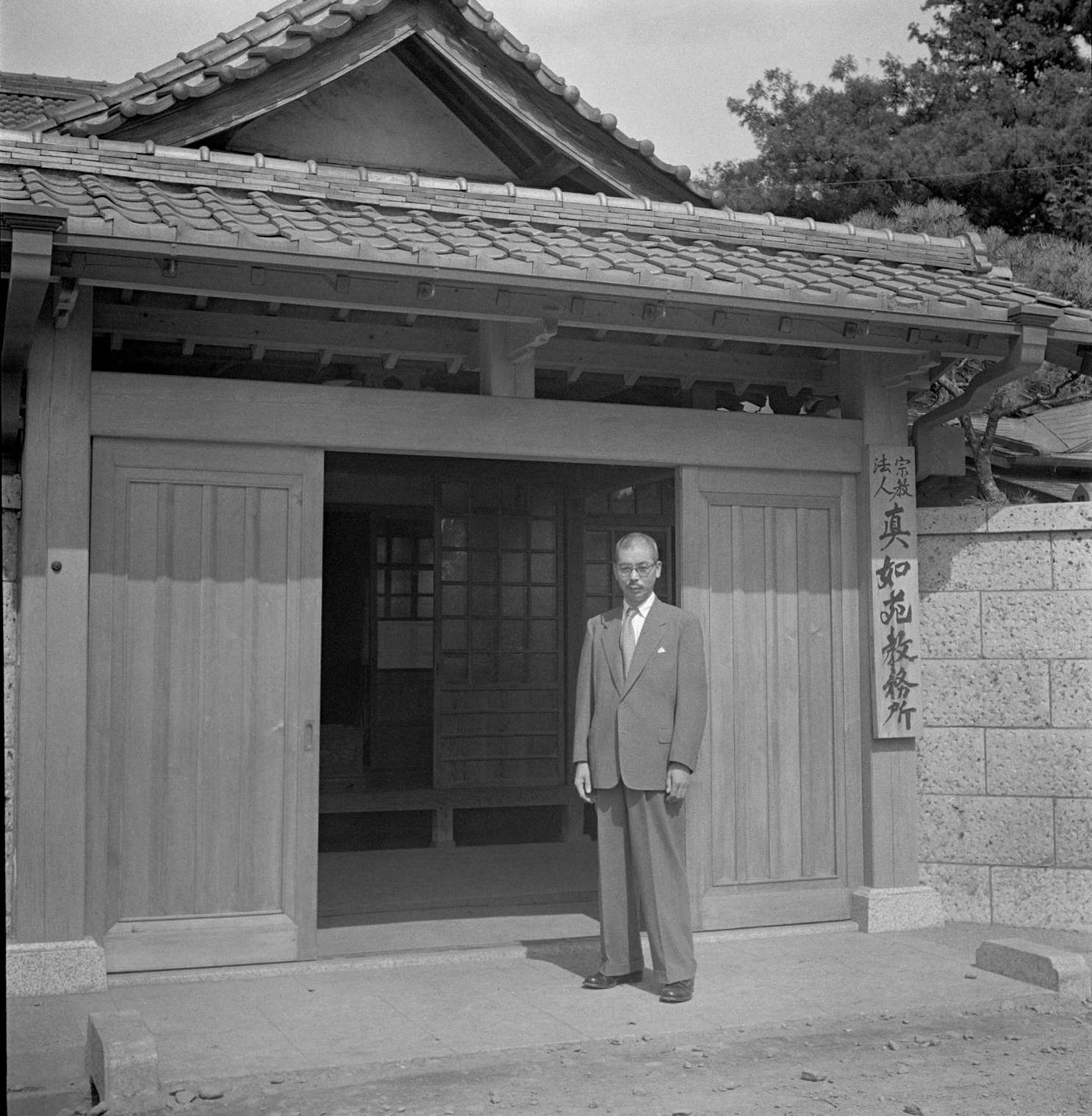 Shinjo, in a suit and glasses, stands at the open door to Shinchoji temple; a plaque with Japanese calligraphy hangs to the right of the door, beneath a tiled roof.