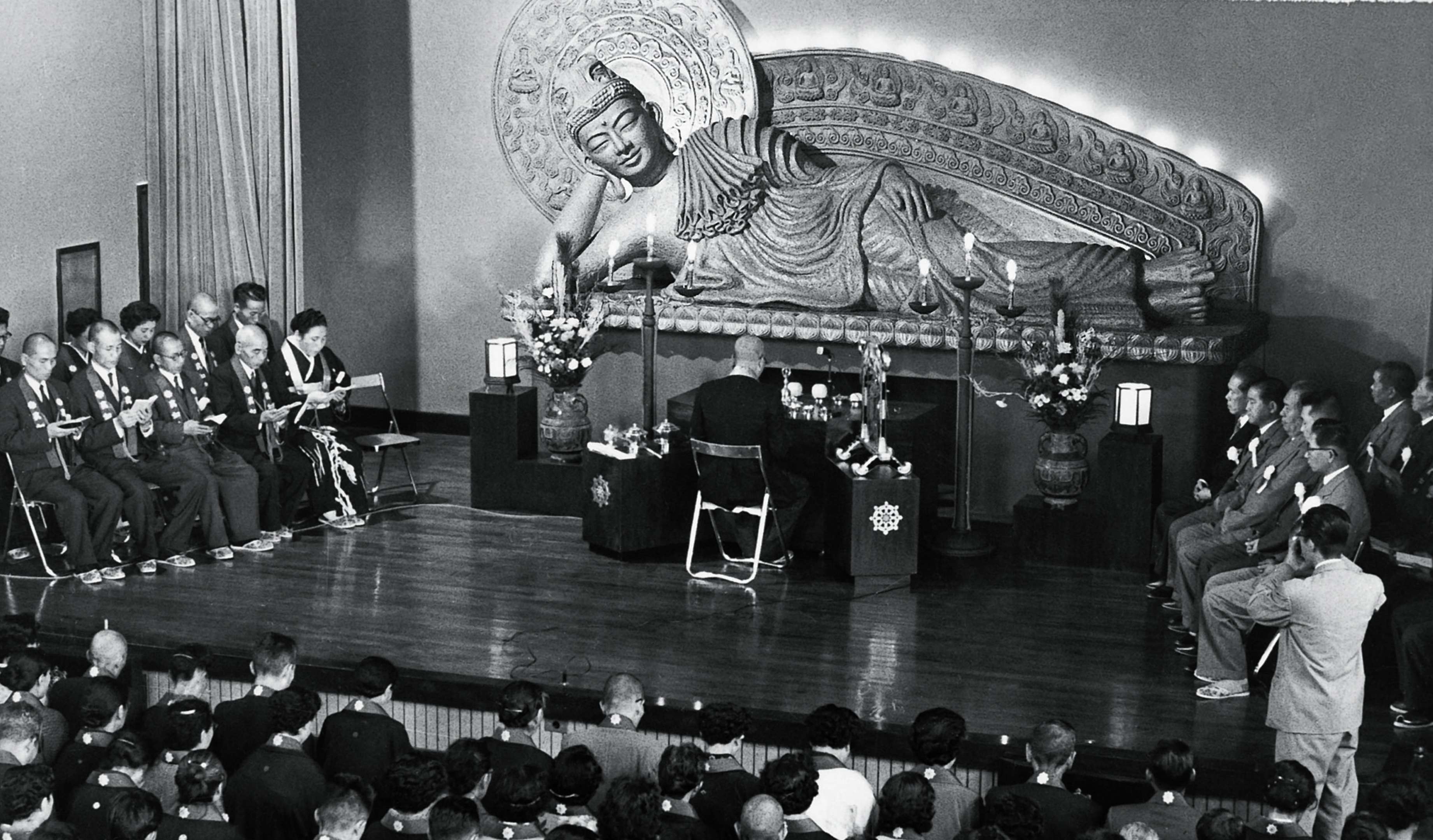 Shinjo, facing away, conducts a rite on a stage before a massive sculpture of a reclining buddha, with a group of priests seated to the left and laypeople to the right, and an audience looking on.