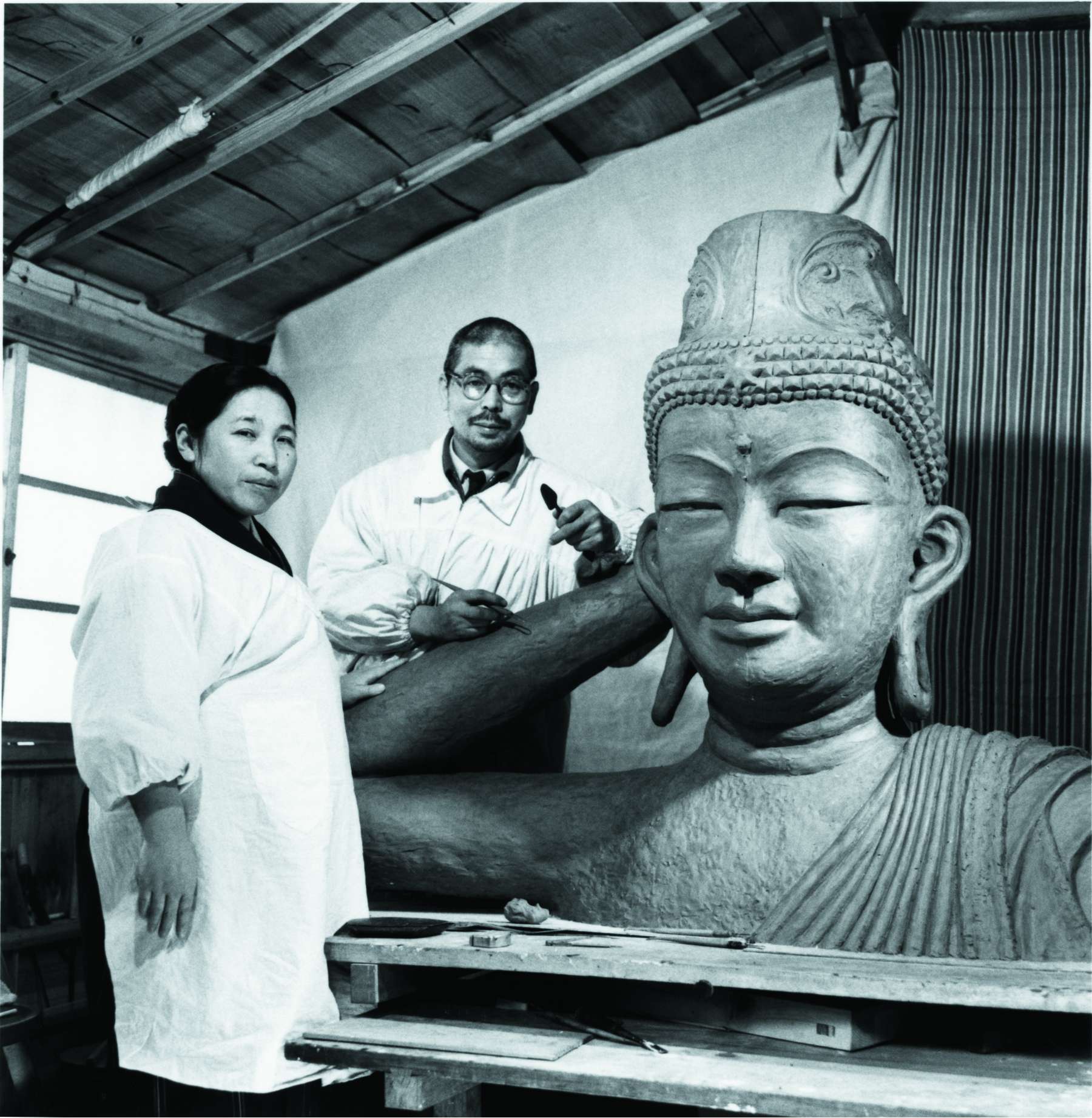 Shinjo, with sculpting implements in hand, and Tomoji, both wearing smocks over their clothing, stand in a studio next to a very large sculpted bust of a smiling buddha.