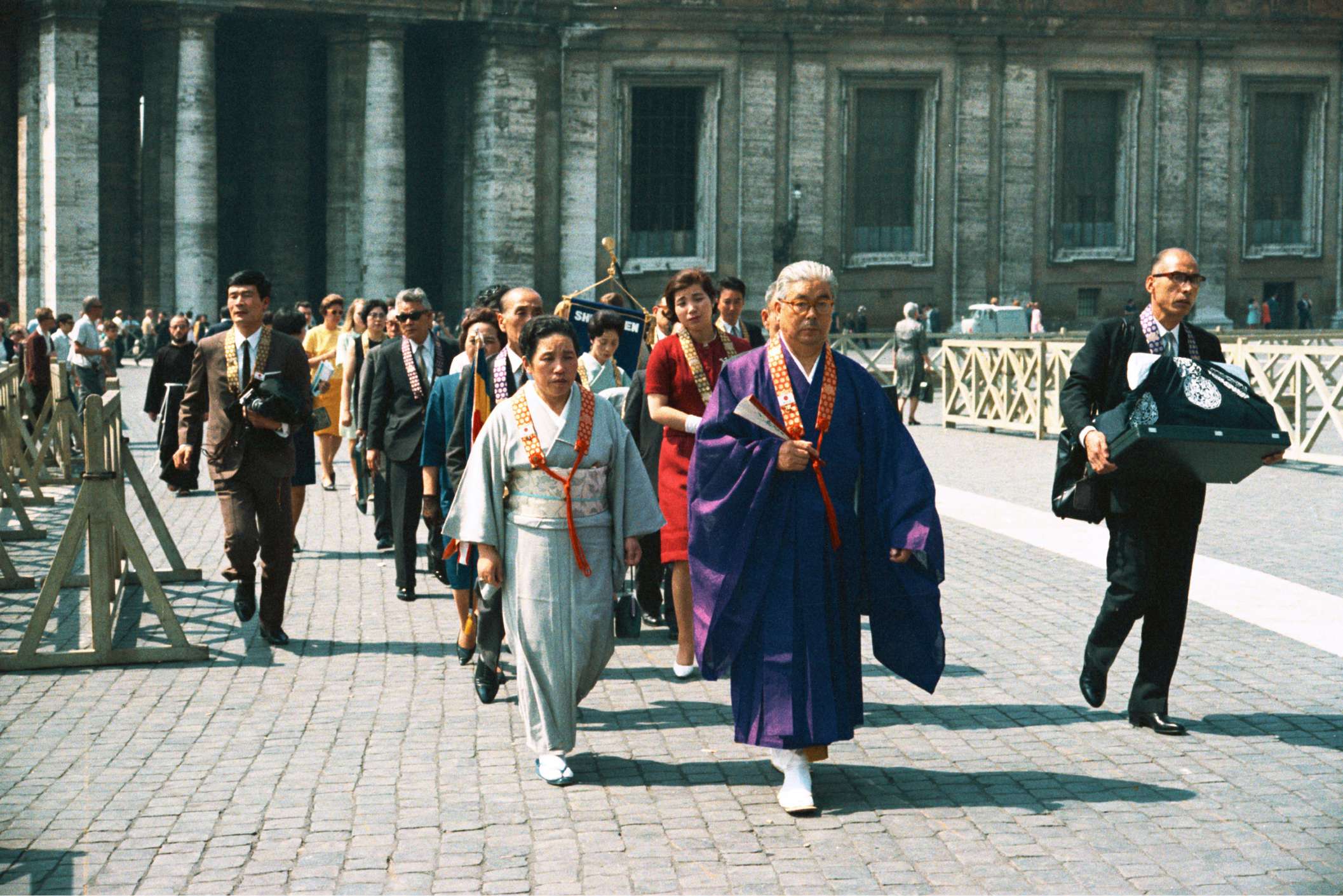Shinjo in purple robes holding a folding fan and Tomoji in a sky blue kimono lead a procession of formally dressed Japanese people through the stone paved, marble pillared courtyard of the Vatican.