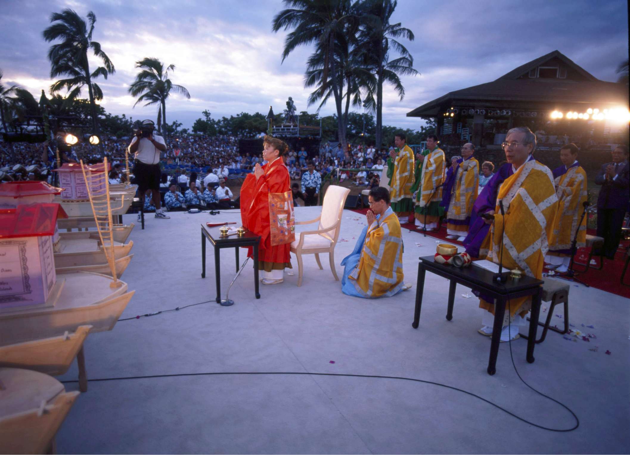 Her Holiness stands in bright orange robes, hands in prayer, on an open-air stage near large boat-like lanterns, with yellow-robed priests behind, and a large audience beneath palm trees looking on.
