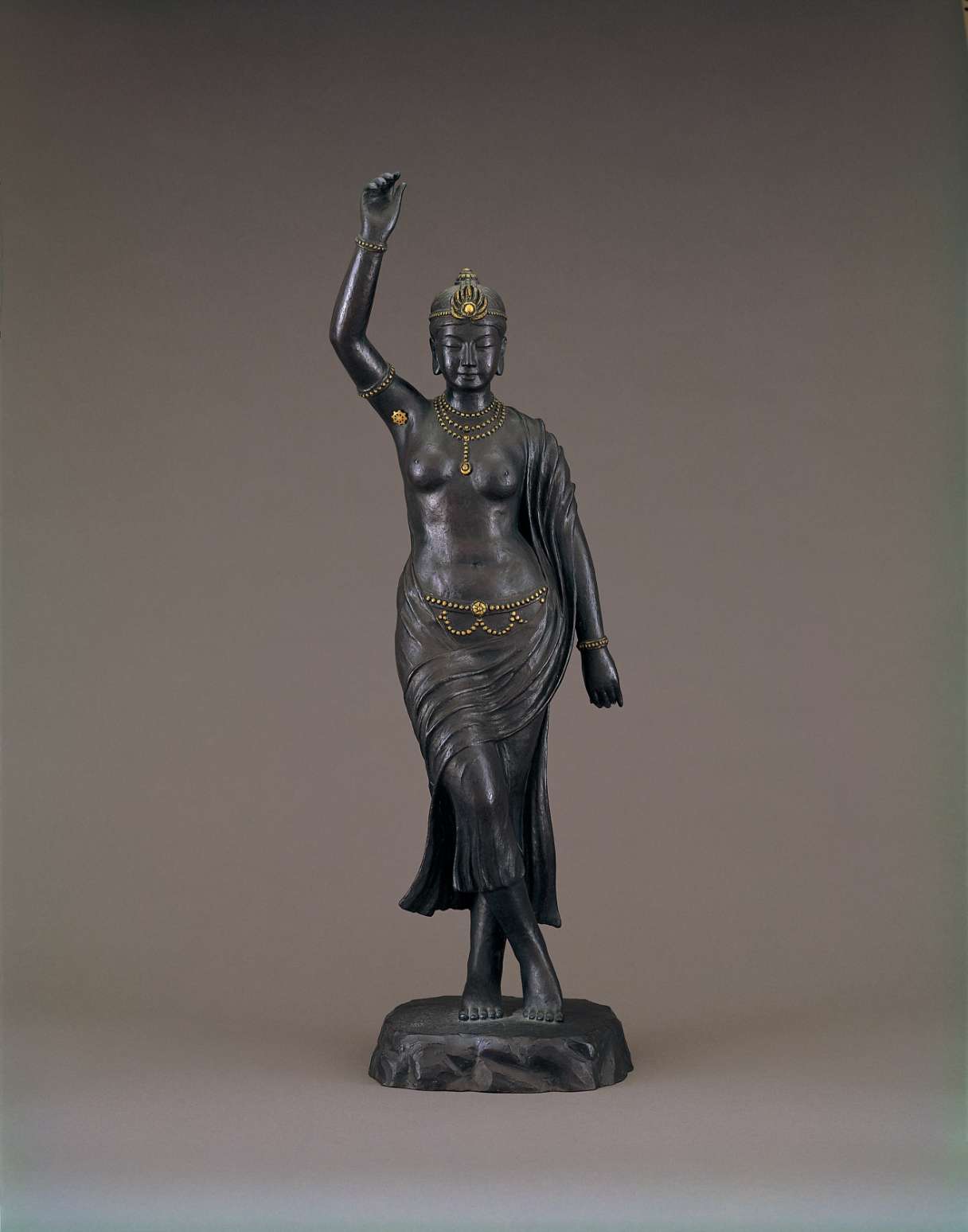 A dark-colored matte statue of a standing woman with serene gaze, adorned with golden jewelry, right arm raised grasping an unseen branch, left at the side, with right leg crossed in front of left.
