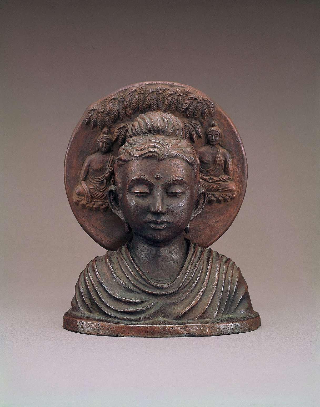 A matte, brown hued bust of Buddha with a plump, youthful face, eyes slightly closed in meditation, backed by a circular nimbus showing a decorative leafy canopy and two seated buddhas.