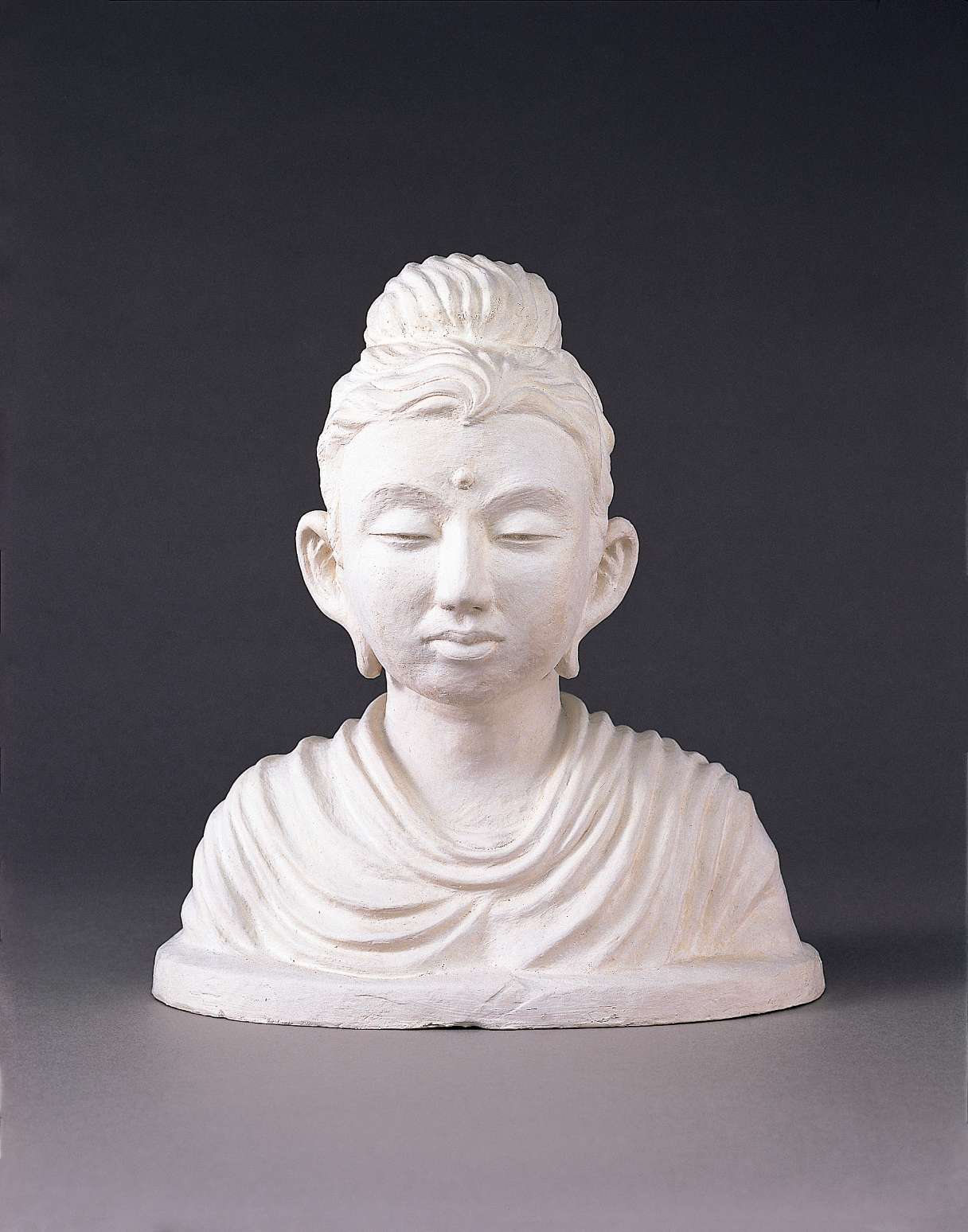 A white plaster bust of Buddha with a plump, youthful face, eyes slightly closed in meditation; the strands of his hair and drapery of his robes form a pleasing wavelike pattern.