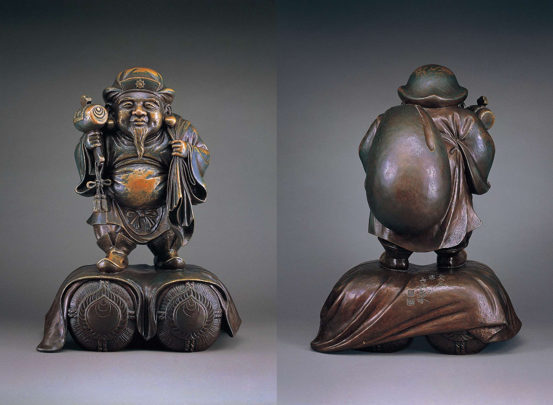 (left) A dark hued statue of a stout, bearded man wearing Chinese tunic, hat, and boots, right hand holding a mallet, left a sack at the shoulder, standing atop two bundles marked with the symbol of jewels. (right) The back of the statue shows the sack, full and bulging with contents, and Japanese calligraphy and and stamps etched into a smooth portion of the statue’s base.