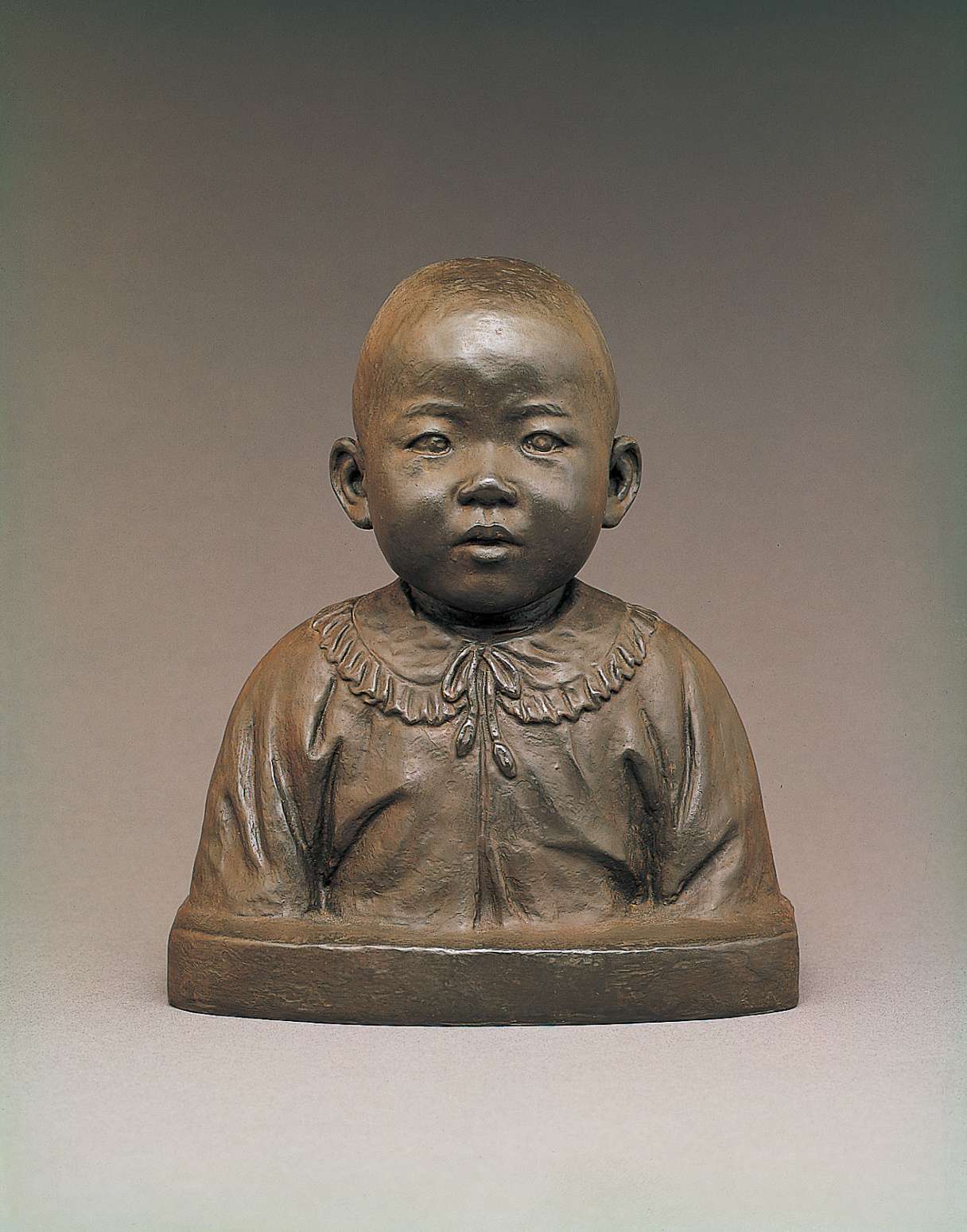 A dark gray bust of a full-cheeked Japanese toddler wearing a frock shirt with a ruffed collar tied at the neck with a bow. The face appears as if gazing expectantly at a parent.