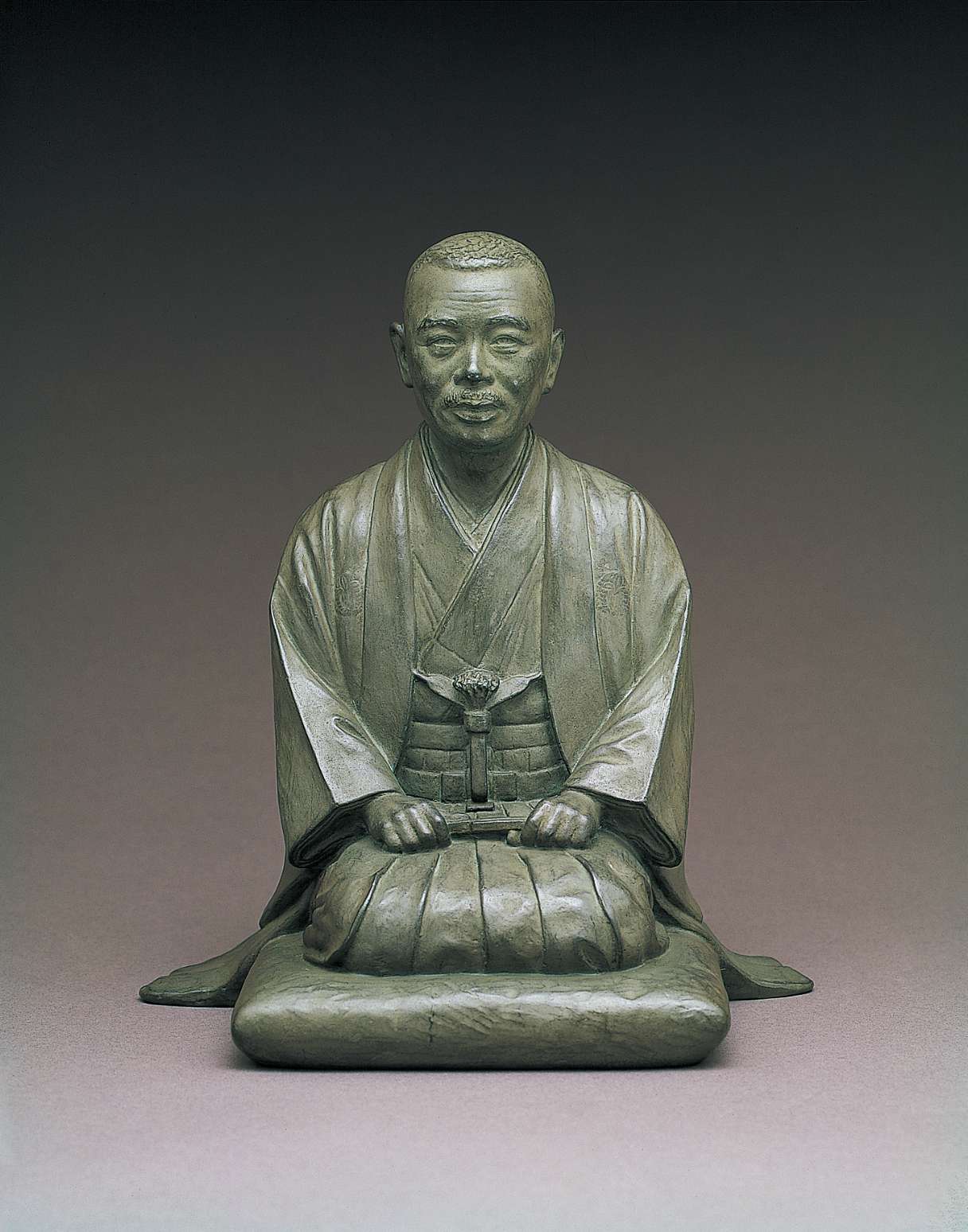 A gray, metallic statue of a late middle-aged Japanese man with close-cropped hair and a mustache wearing traditional Japanese kimono, kneeling on a cushion holding a folding fan in his lap.