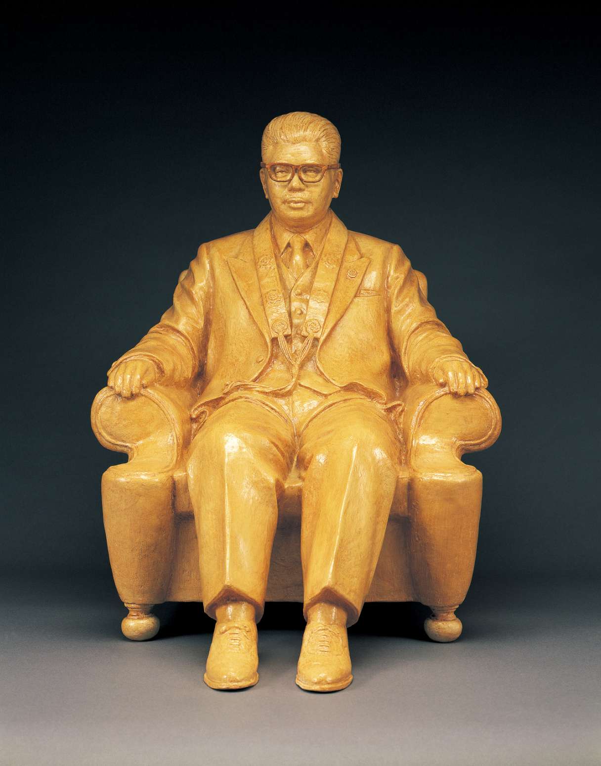 An ochre hued statue of a spectacled, mustachioed middle-aged Japanese man in suit and tie, full head of hair, seated in an upholstered chair, his arms resting on the armrests, feet flat on the floor.