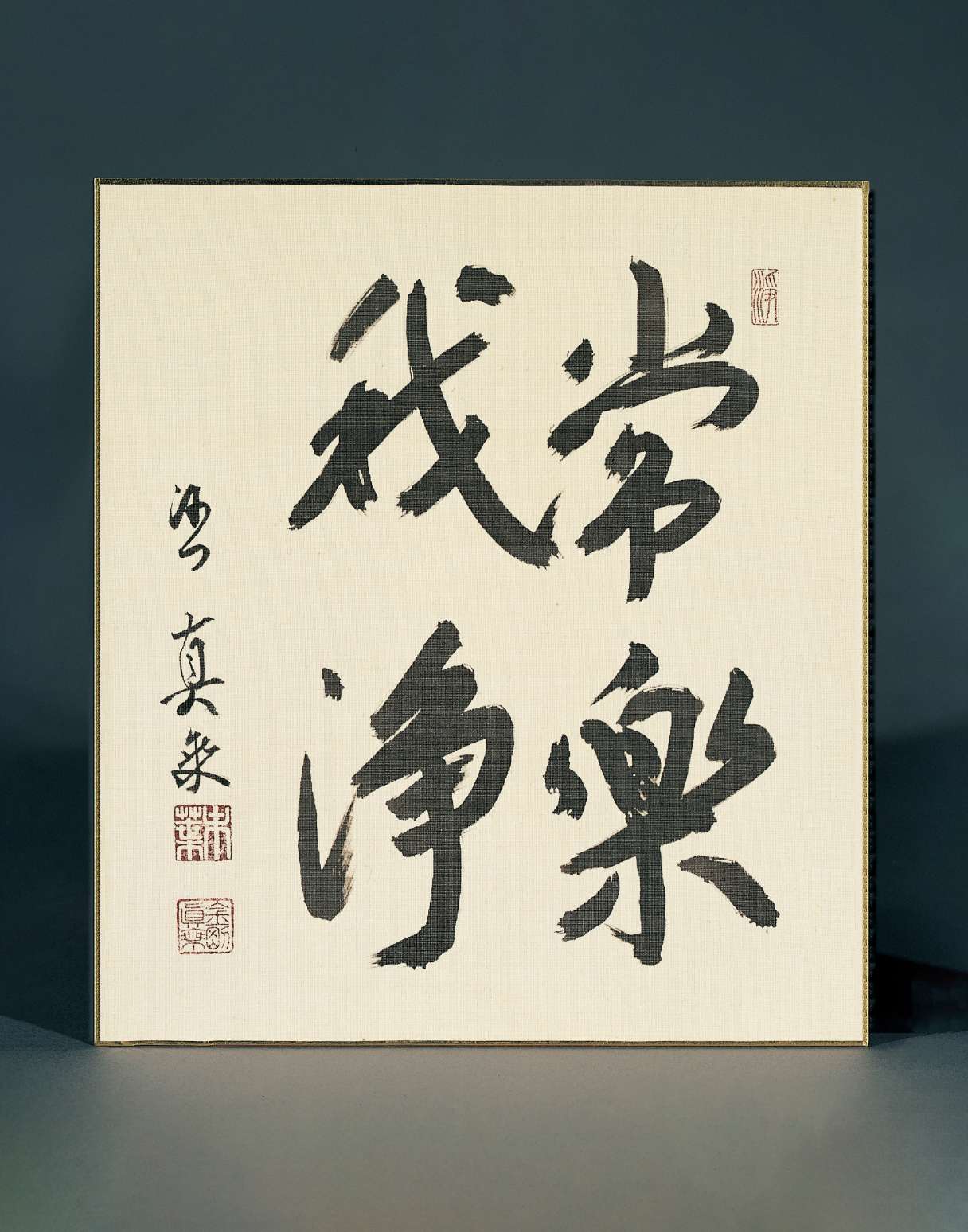 Four large Japanese calligraphy characters are written with brush on a squat, rectangular sheet of paper; to the left another smaller line of calligraphy is written with two stamp impressions in black.