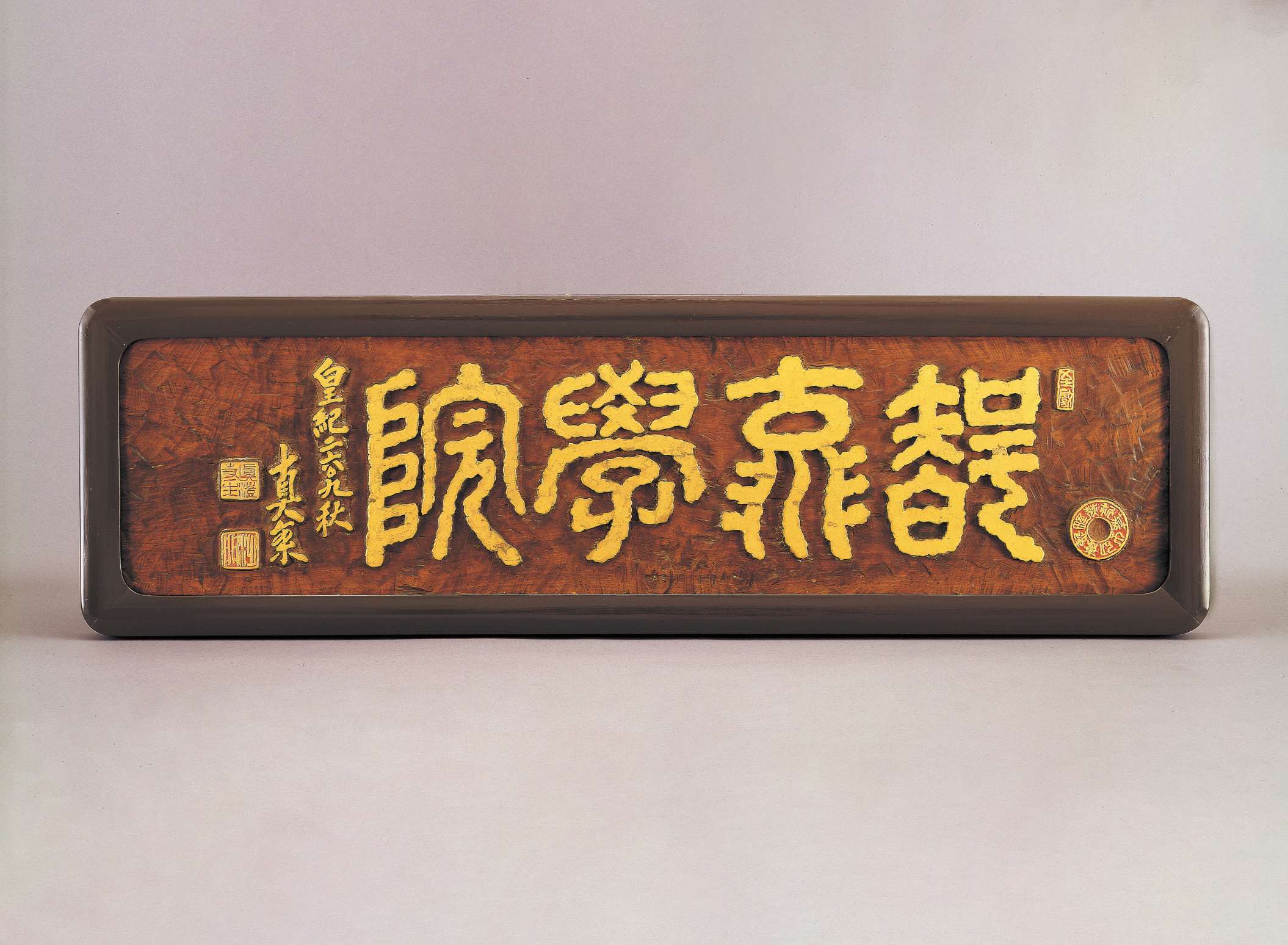 Stylized, thick-lined Japanese characters in gold are carved into a slab of cherry stained wood, framed by a polished border, embellished by adjacent golden calligraphy and square seals.