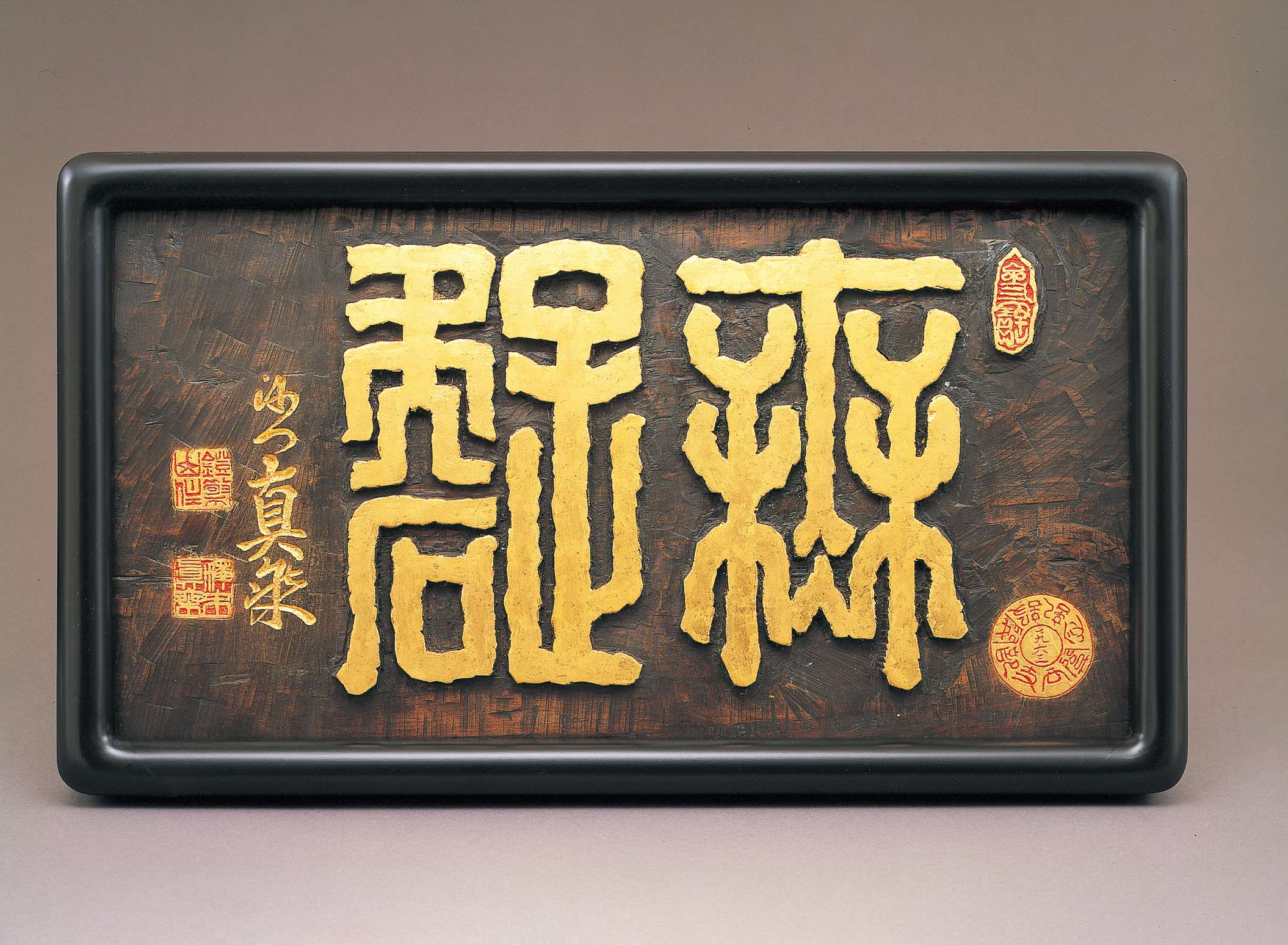 Stylized, thick-lined Japanese characters in gold are carved into a slab of dark brown wood in a black frame, embellished by adjacent golden calligraphy and red and gold seals.