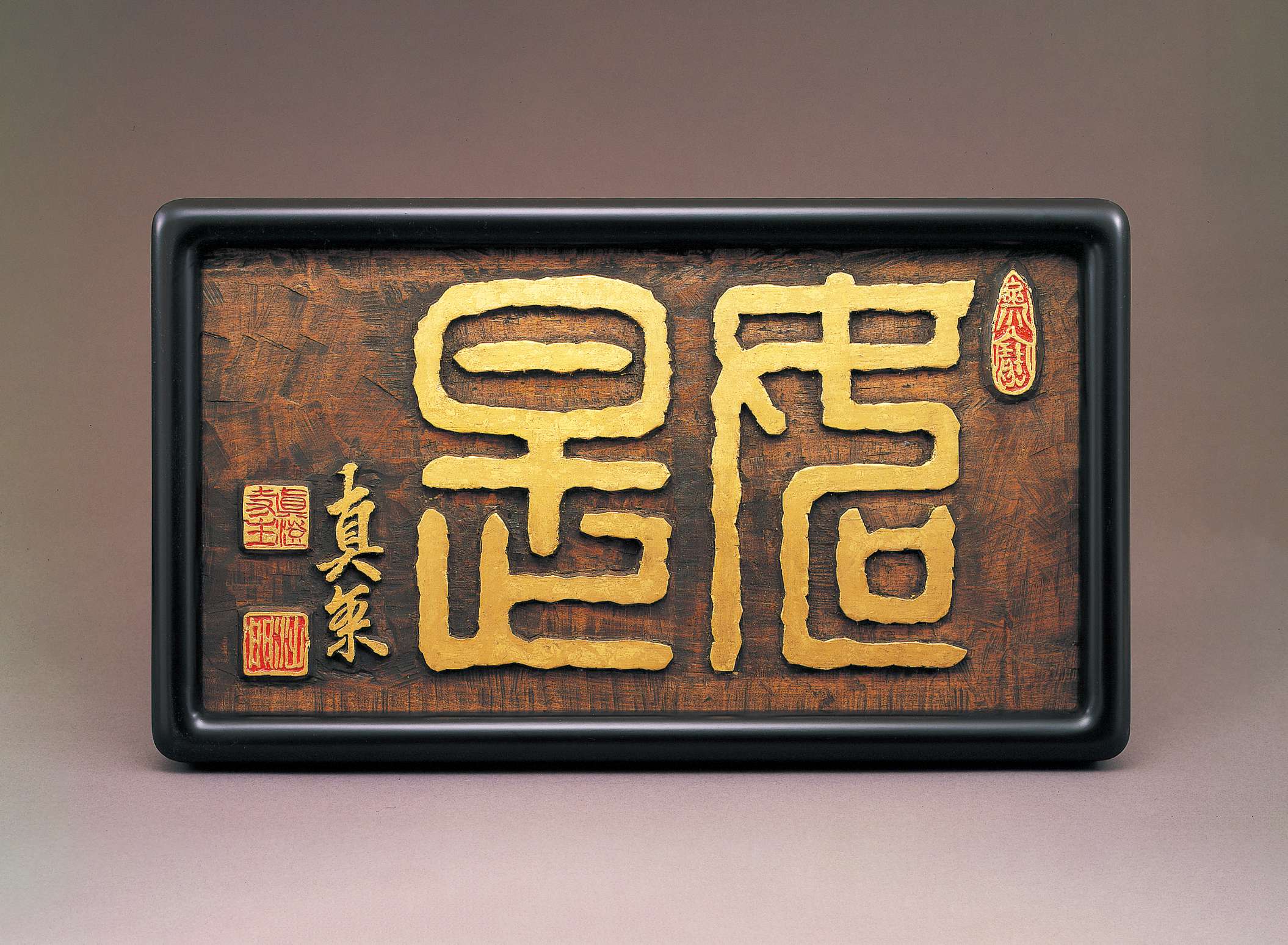 Stylized, thick-lined Japanese characters in gold are carved into a slab of dark brown wood in a black frame, embellished by adjacent golden calligraphy and red and gold seals.