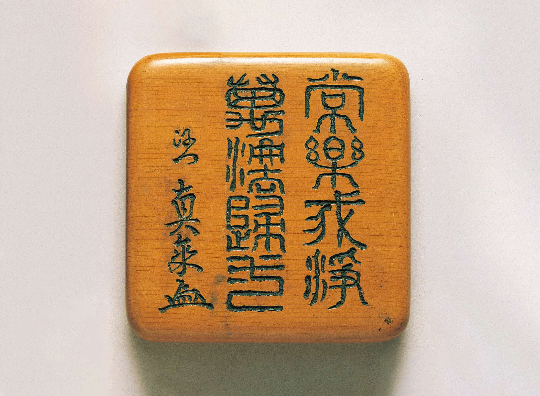 A square slab of smooth, polished sienna colored wood is engraved with vertically oriented, stylized Japanese characters, to the left of which is engraved a line of flowing Japanese calligraphy.