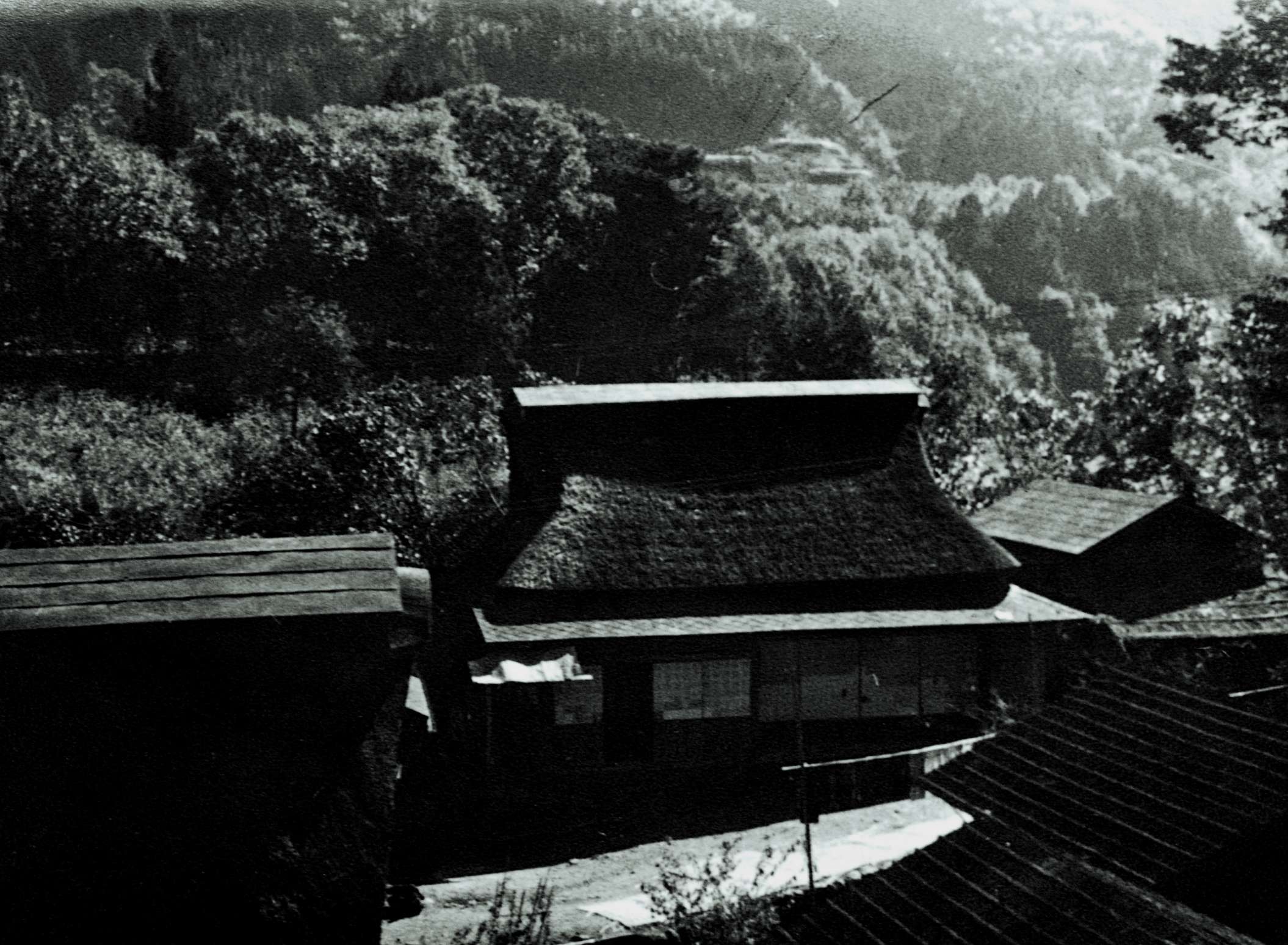 A black and white photo of an idyllic Japanese village, with a large thatched roof house in the center; early morning sunlight casts shadows and bathes distant mountain groves in a haze of light.