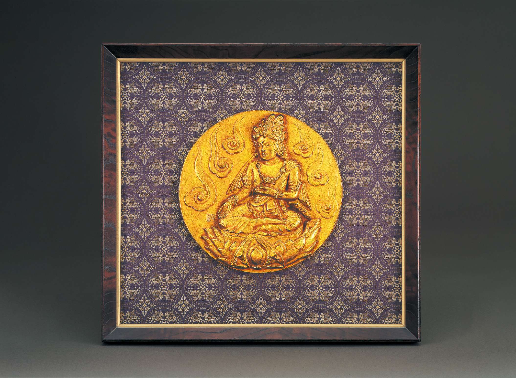 A circular golden relief of a bodhisattva in royal robes and jewelry, hair in topknot, sitting cross-legged atop a lotus seat, right hand resting palm upturned on knee, left hand raised, upturned.